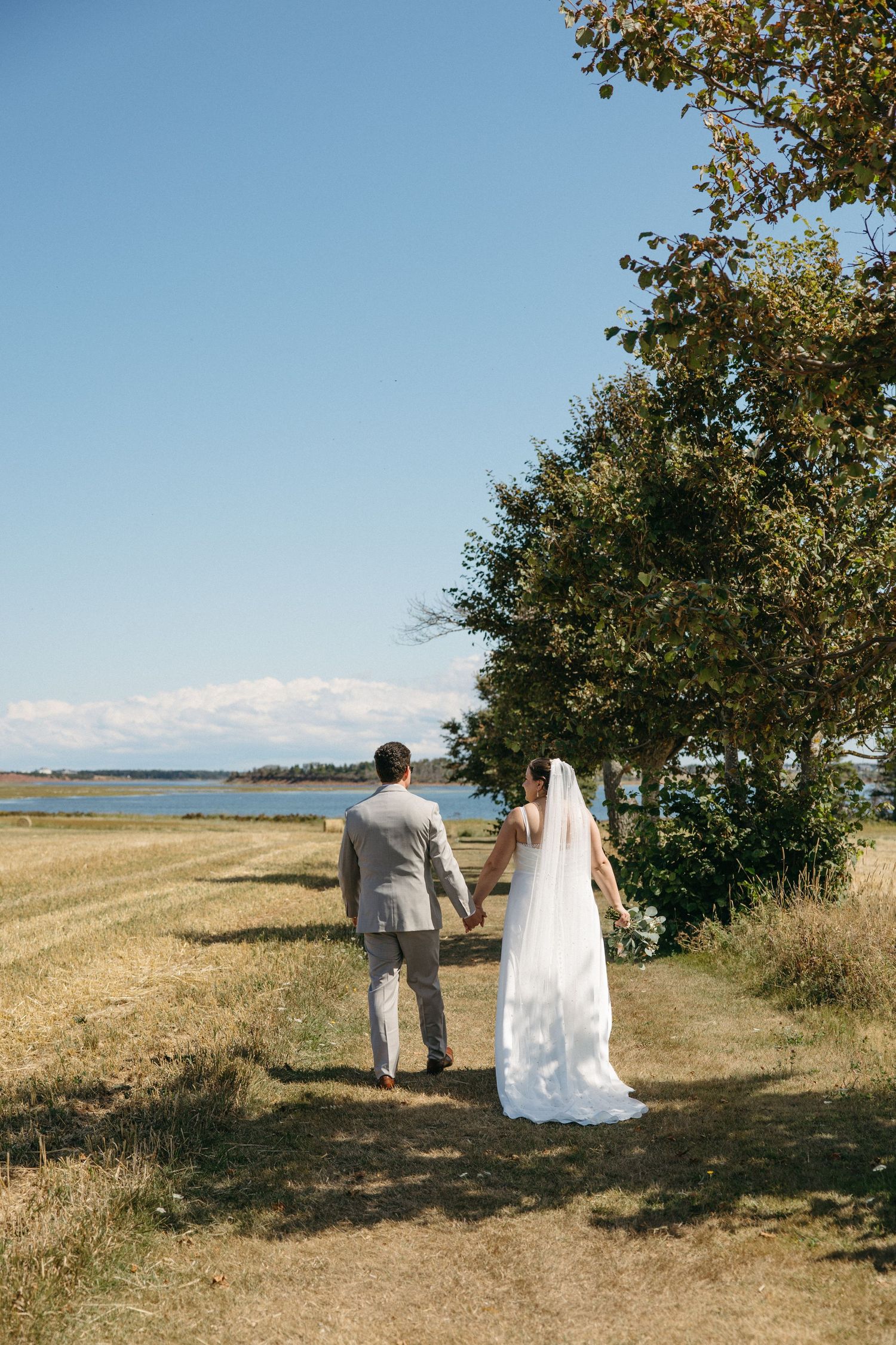 Bride and groom walk hand in hand through golden fields toward a serene waterfront on their wedding day.