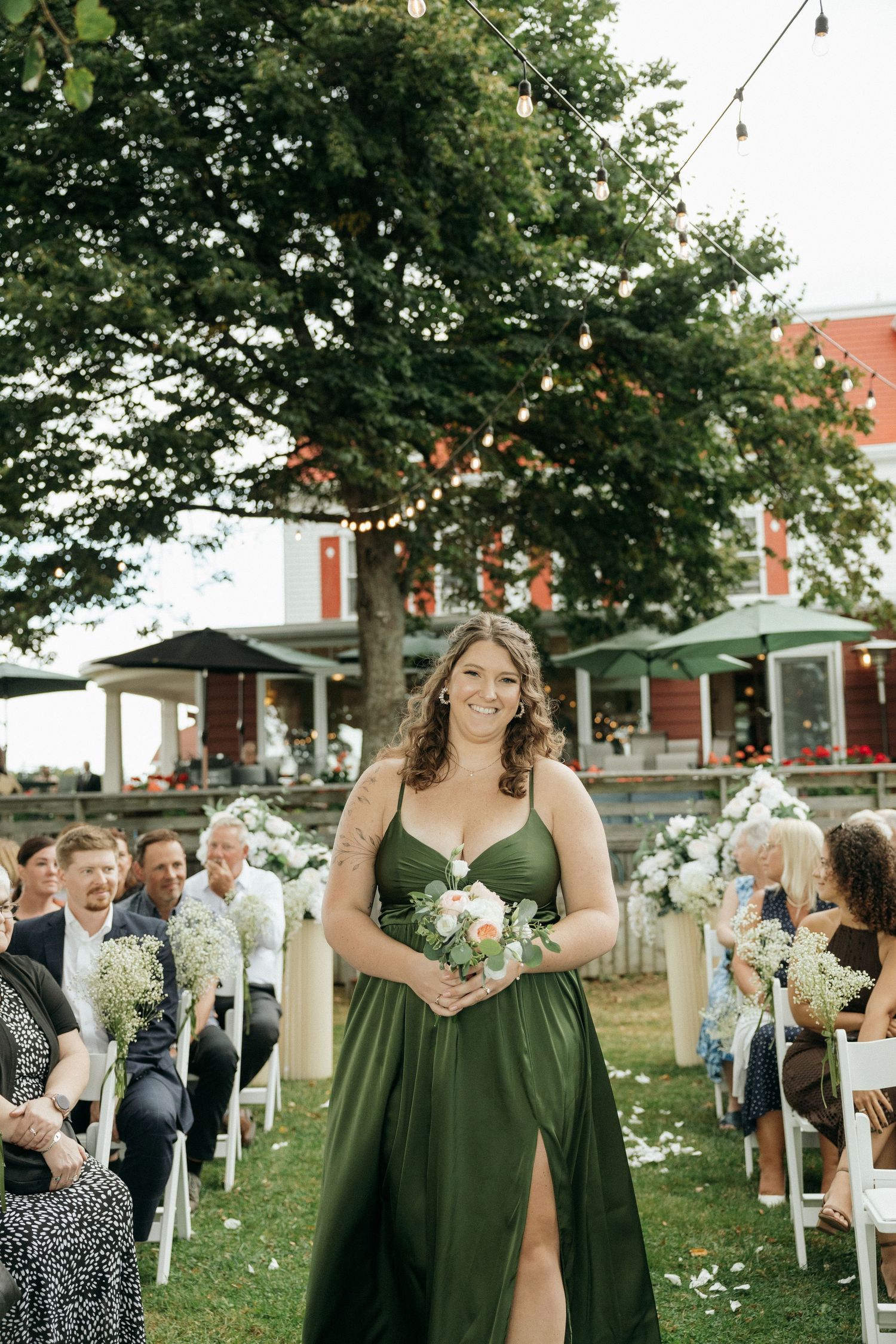 Bridesmaid in green satin dress walking down outdoor wedding aisle holding bouquet.