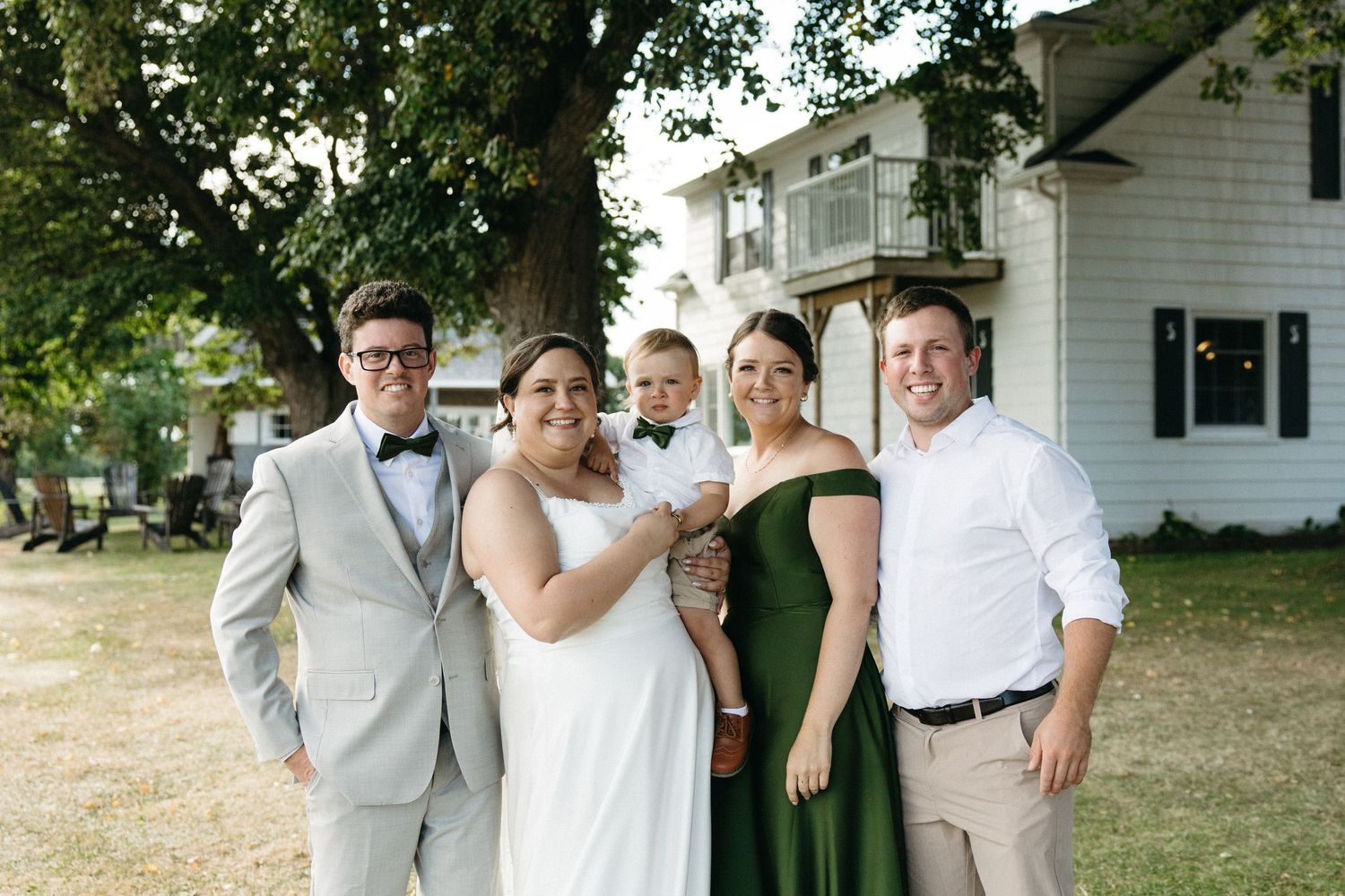 Wedding party group portrait with bride, groom, bridesmaid in green dress, and guests outdoors.