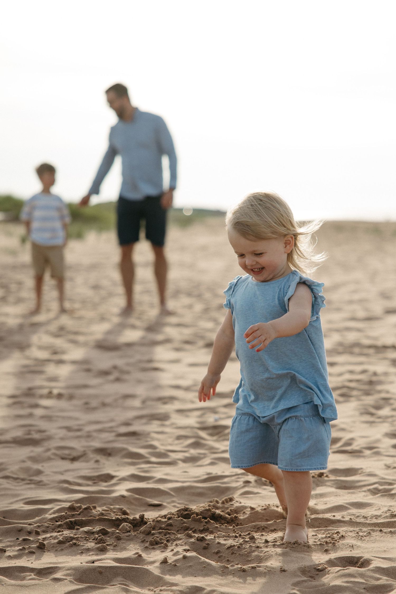 Smiling toddler girl in blue outfit walking on sandy beach with father and boy behind her.