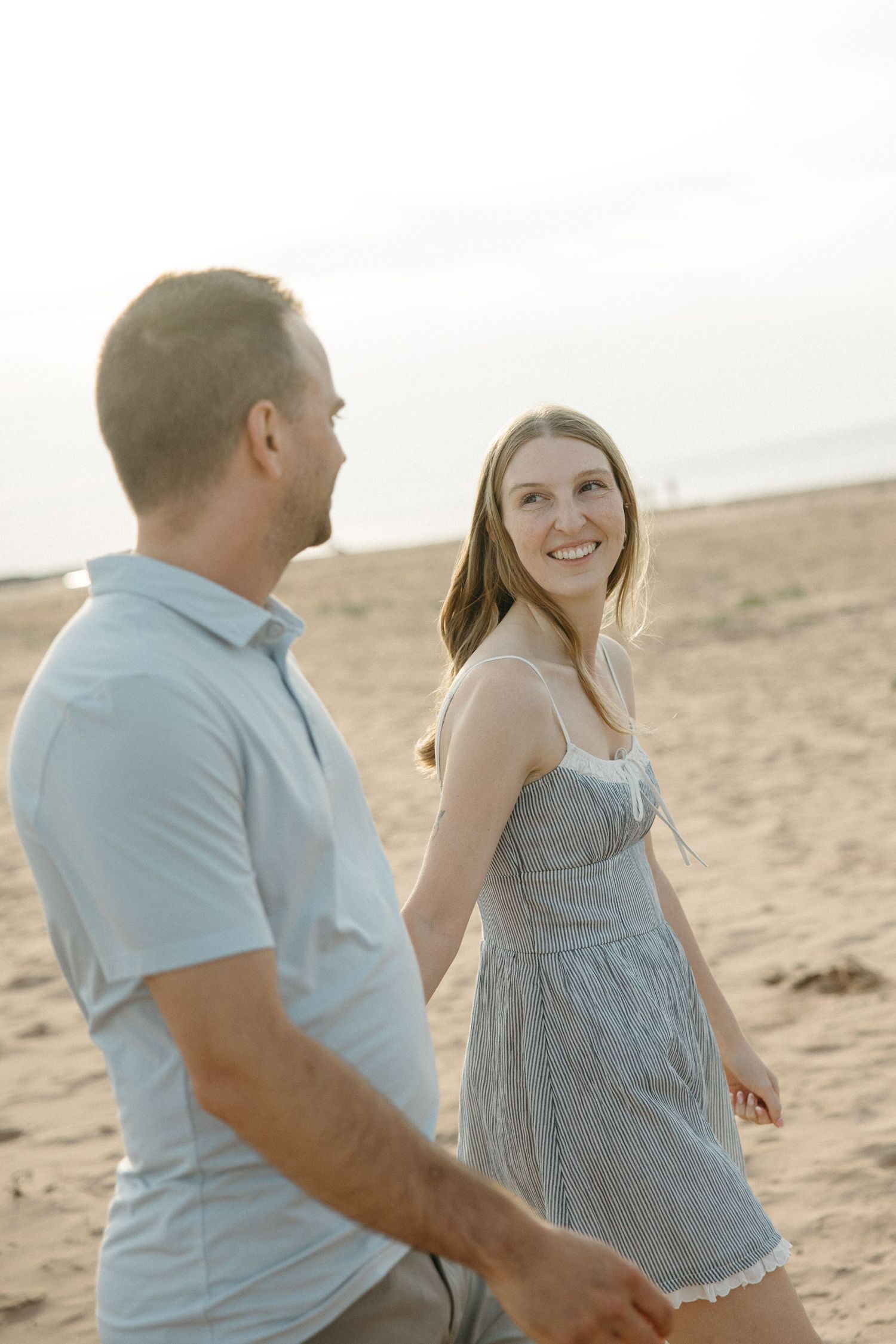 A smiling couple walks hand in hand on a sunny sandy beach, the woman wearing a blue sundress.