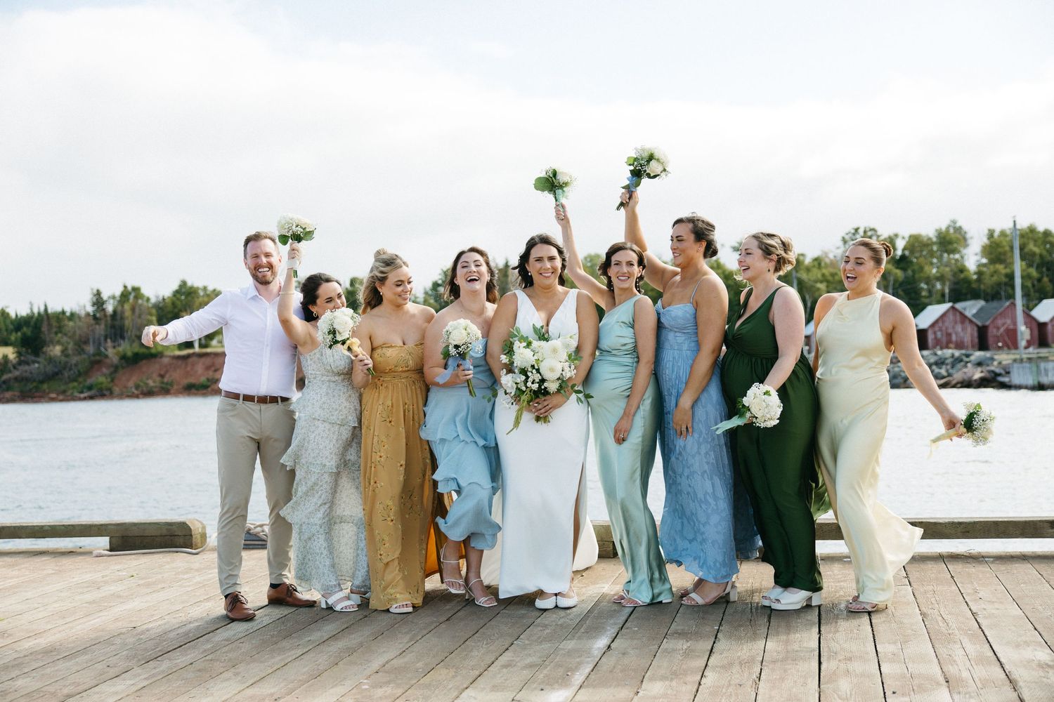 Wedding party members in light blue and white dresses stand together on a wooden dock by the water.