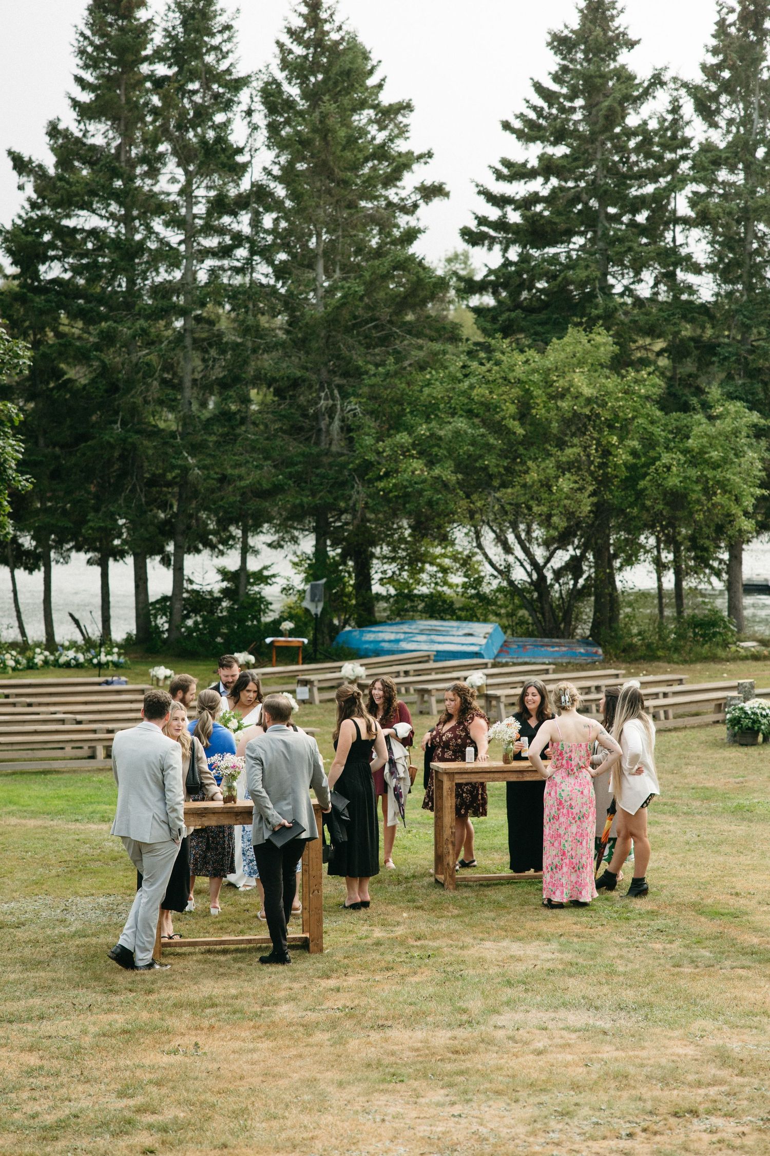 Wedding guests gather around wooden tables on a grassy lawn with tall evergreen trees in the background.