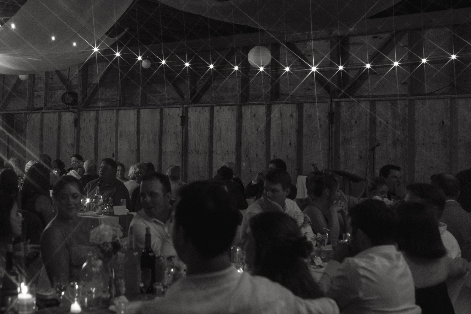 Dimly lit barn reception with string lights and guests gathered at tables.