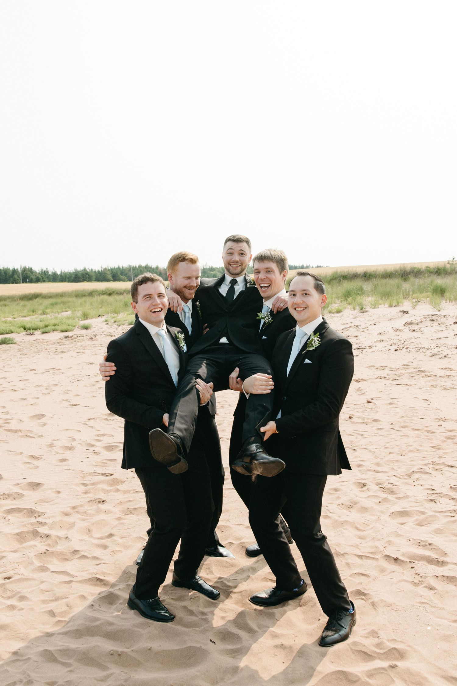 Groomsmen in black suits pose playfully on sandy beach.