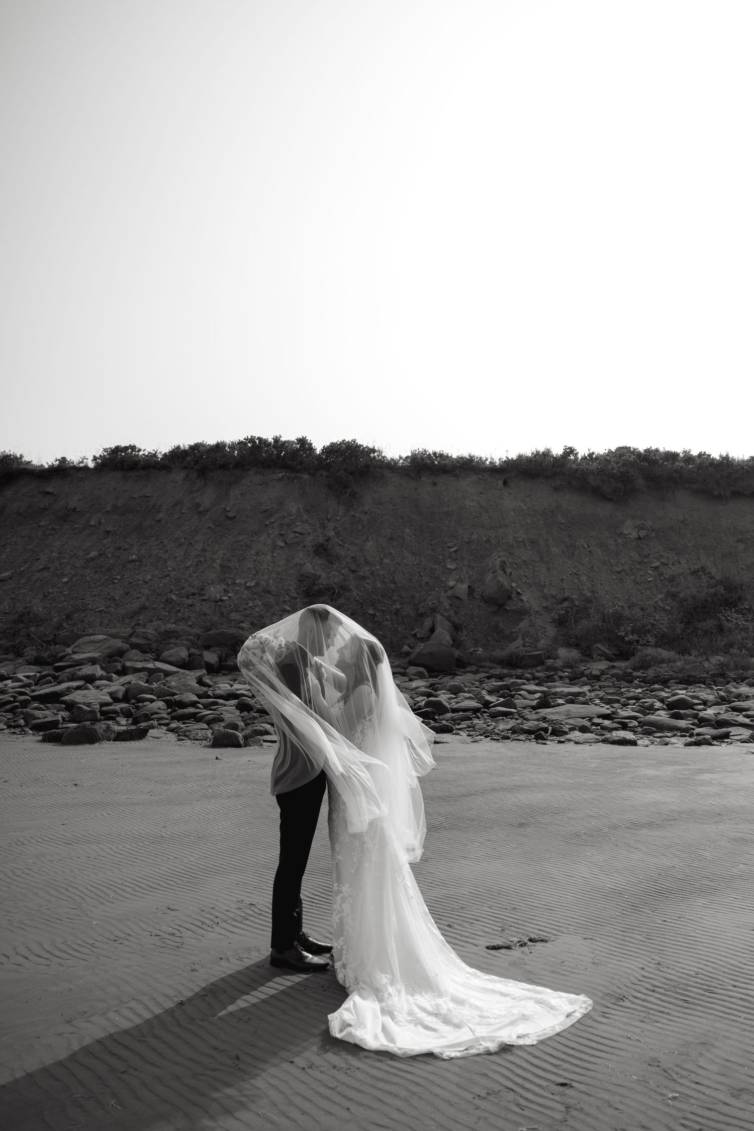 A couple shares an intimate moment on a beach as the bride's veil flows in the wind.