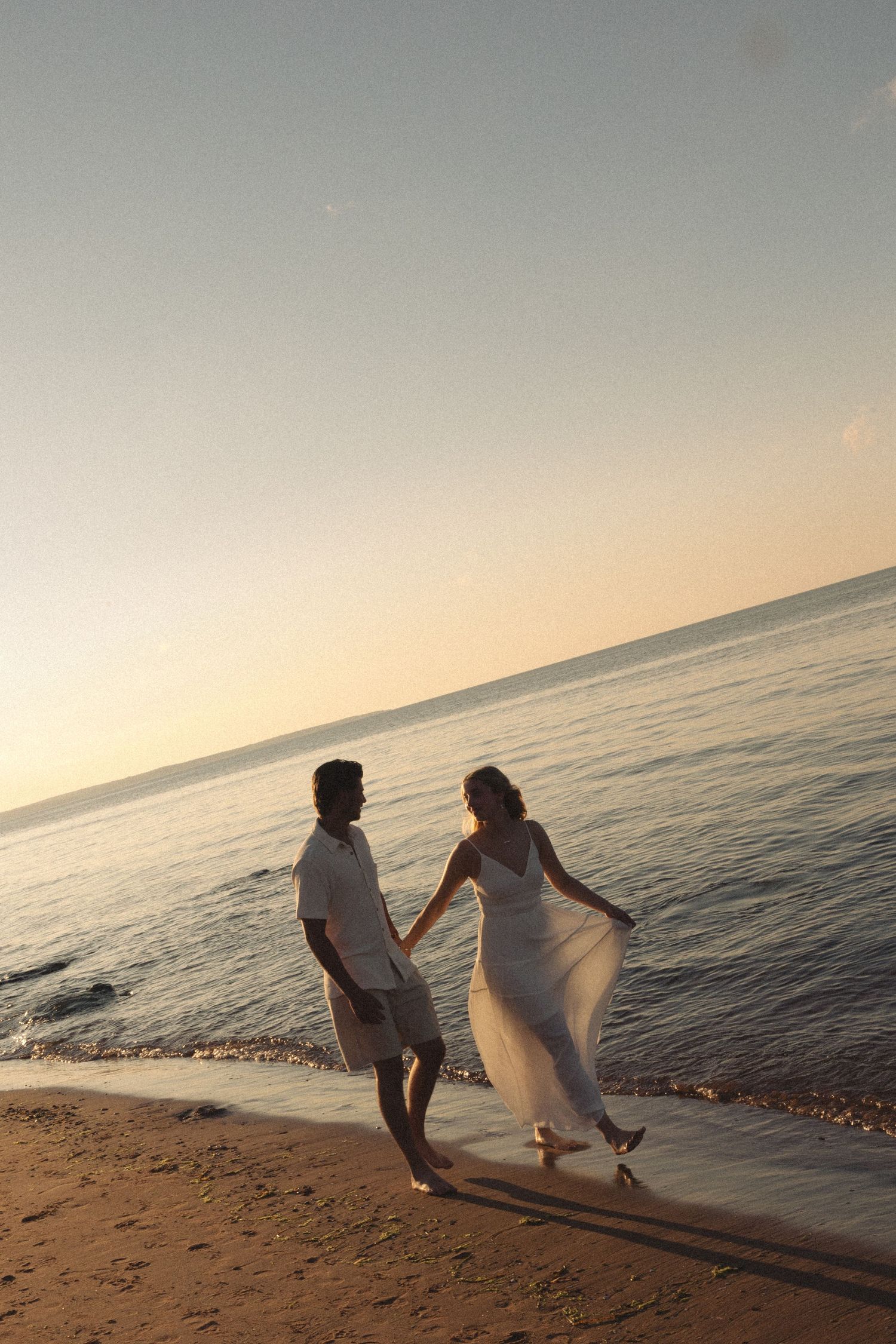 Couple in white attire holding hands walking along sandy beach at golden sunset.
