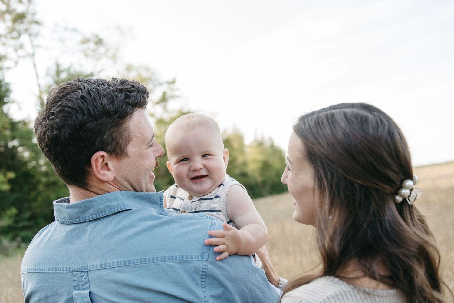 Happy baby smiling at parents during outdoor family photo session in a sunny golden field.