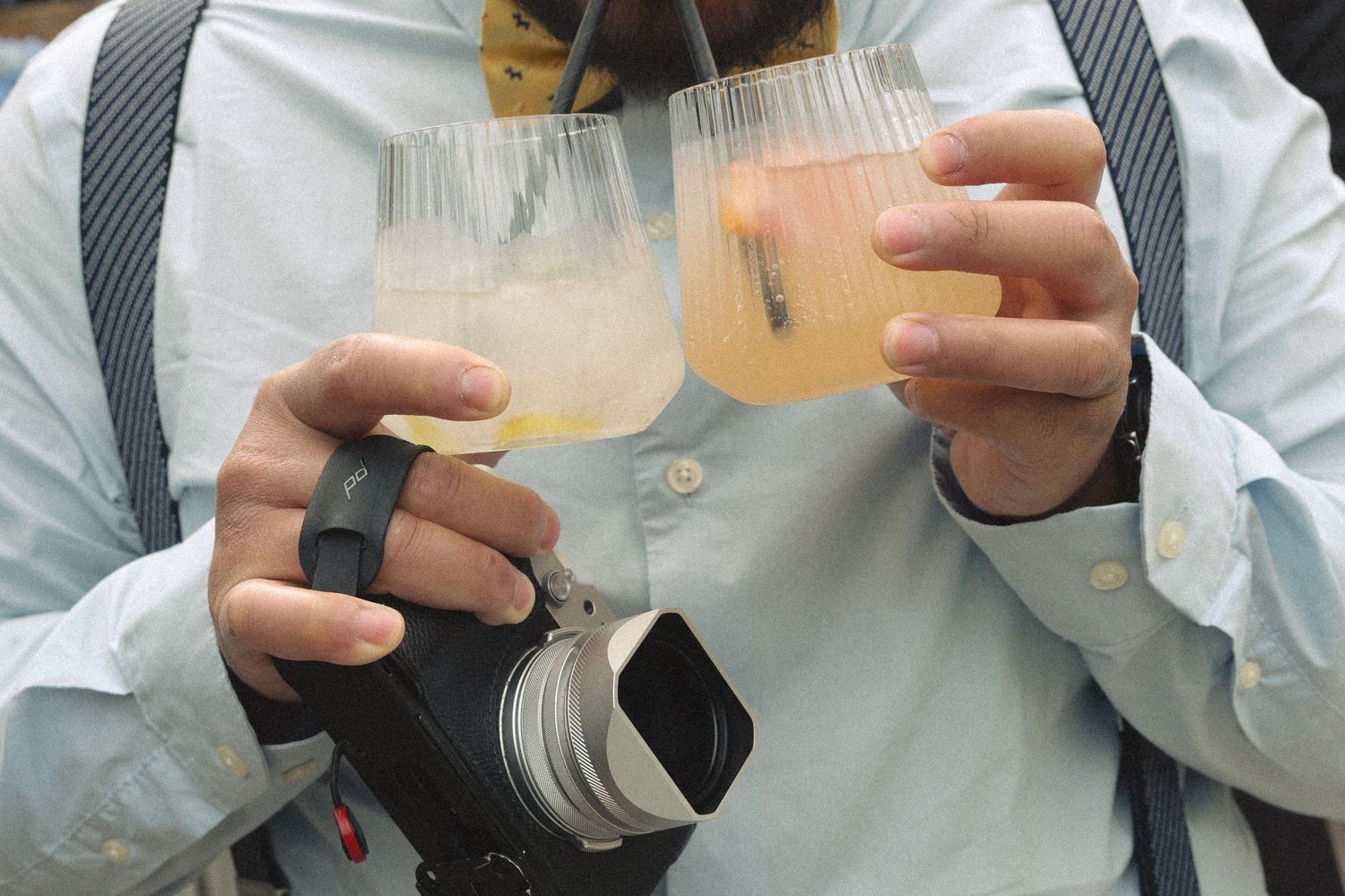 Wedding photographer holding two stemless glasses of pale cocktails while wearing a camera.