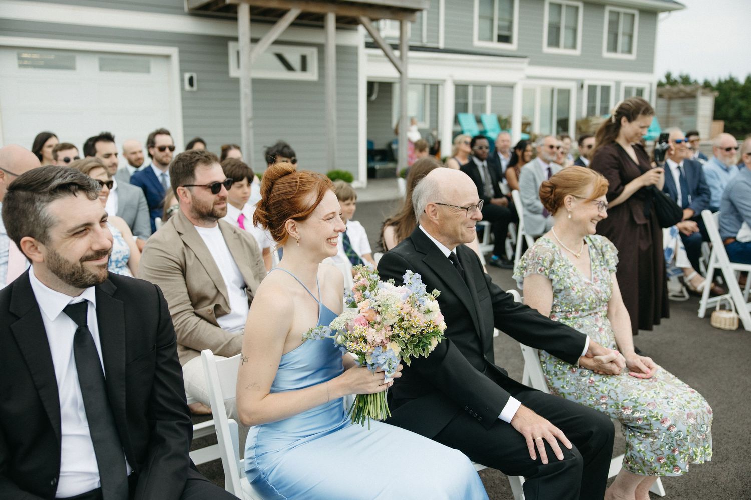 Wedding guests seated at outdoor ceremony, bridesmaid in blue dress holding colorful floral bouquet.