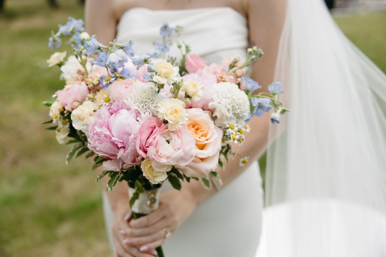 Bride holds lush bouquet of pink peonies, peach roses, and blue flowers in white gown.