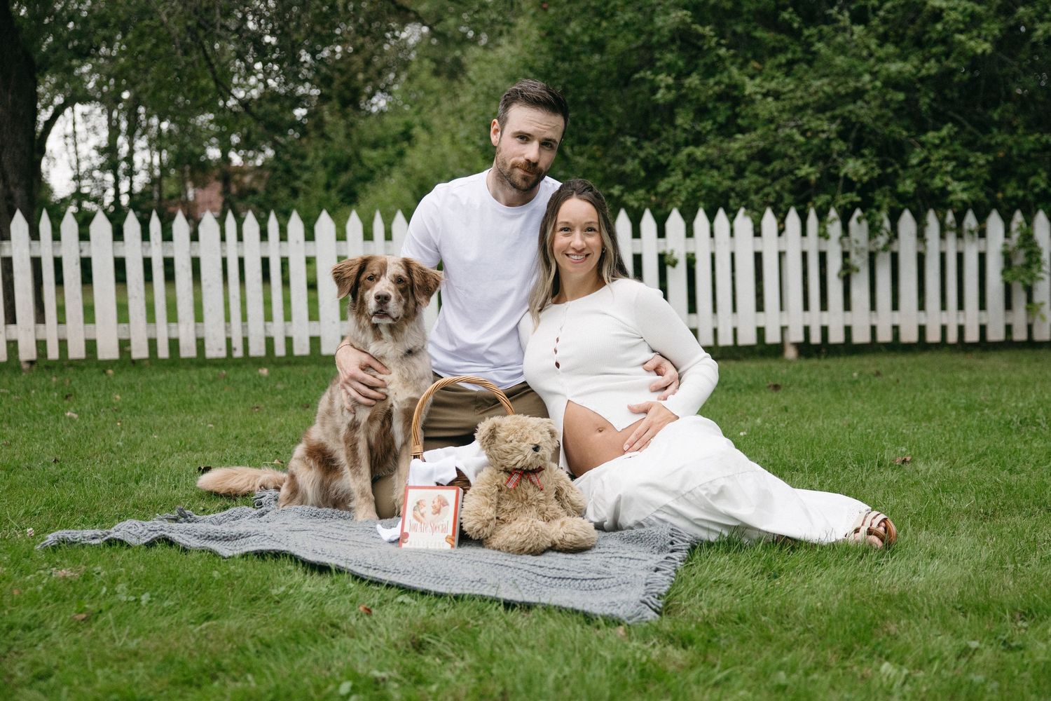Pregnant couple sits on blanket in garden with brown dog and teddy bear, white picket fence background.