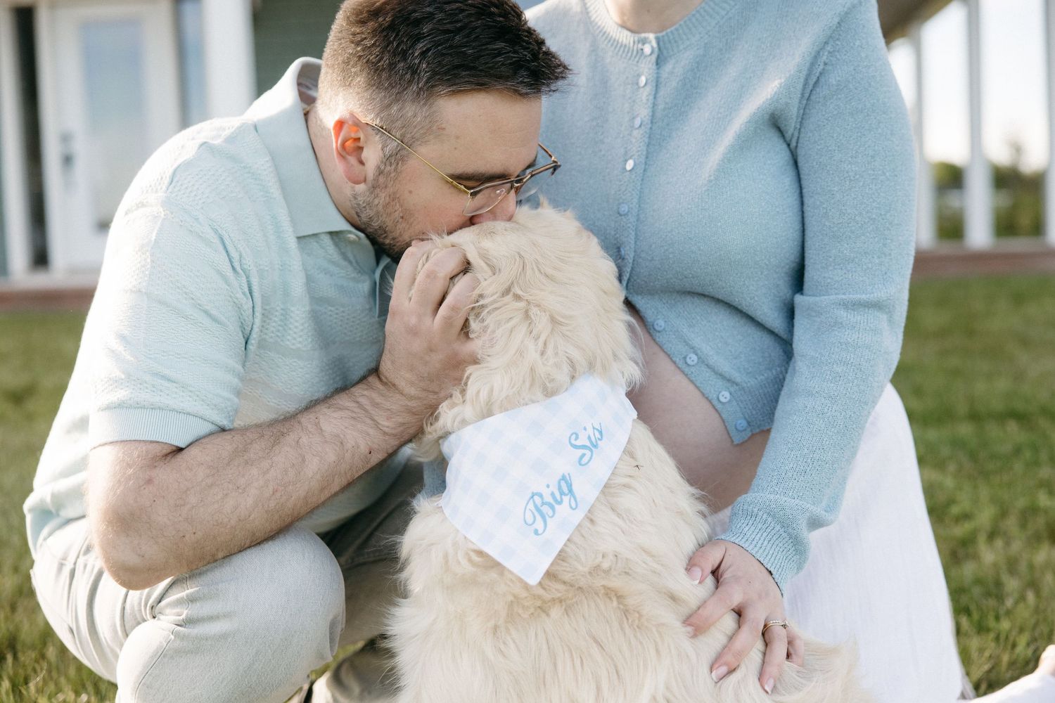 Couple kissing their fluffy golden doodle dog wearing a Big Sis bandana outdoors on green grass.