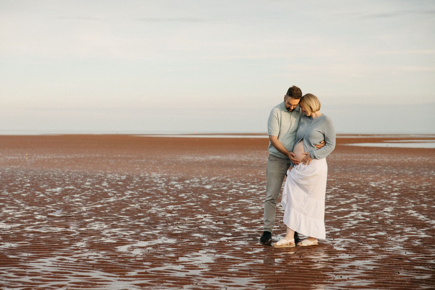 Couple embracing on a red sand beach at low tide, expecting parents sharing a tender moment by the water.