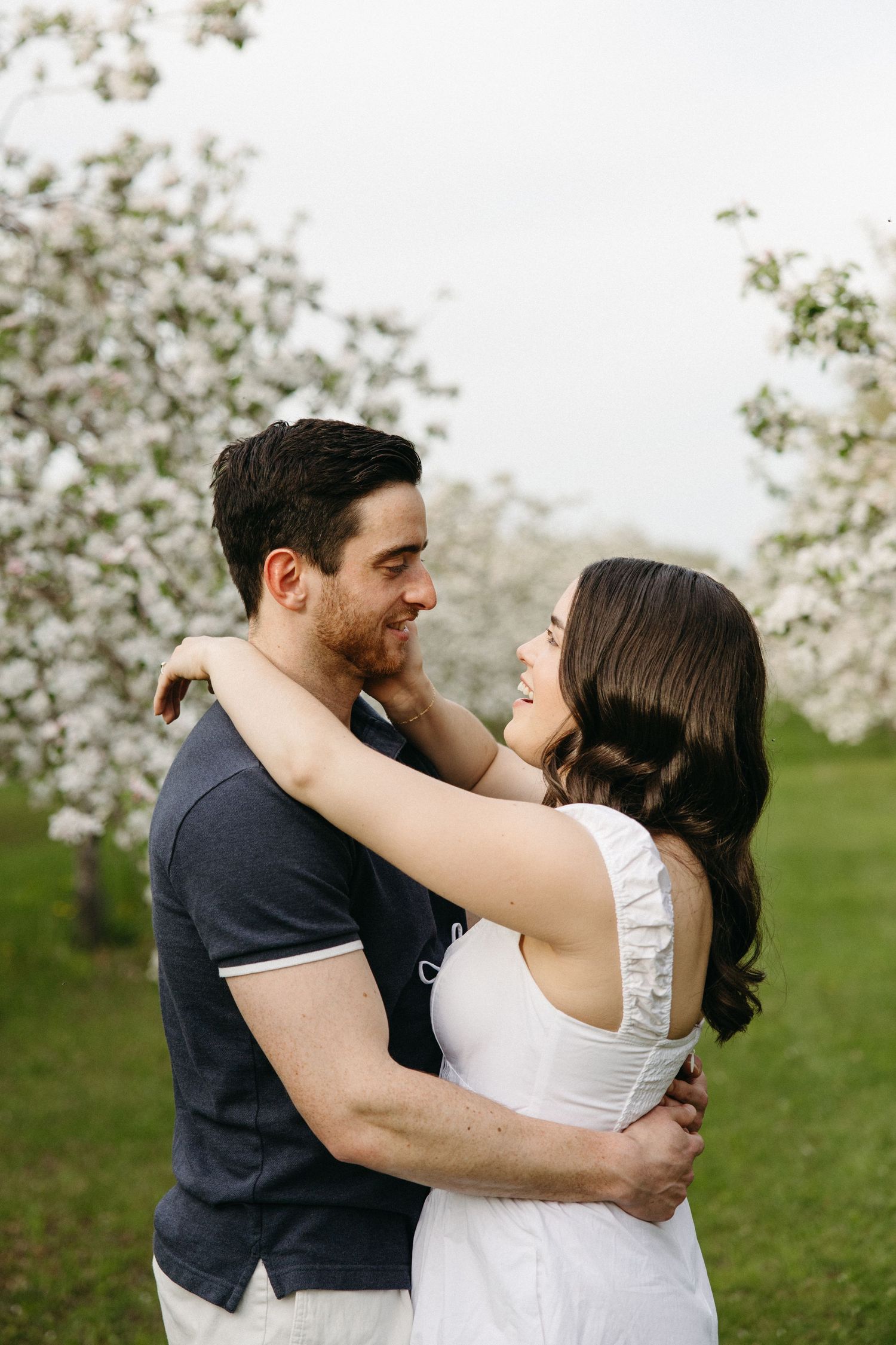 A couple shares a tender embrace and loving gazes surrounded by white blooming trees.