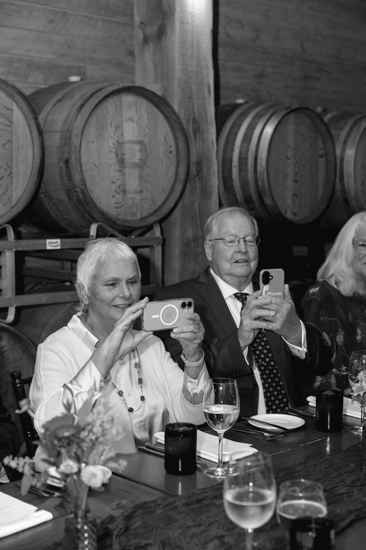 Black and white photo of guests dining and socializing in a rustic wine cellar with wooden barrels.