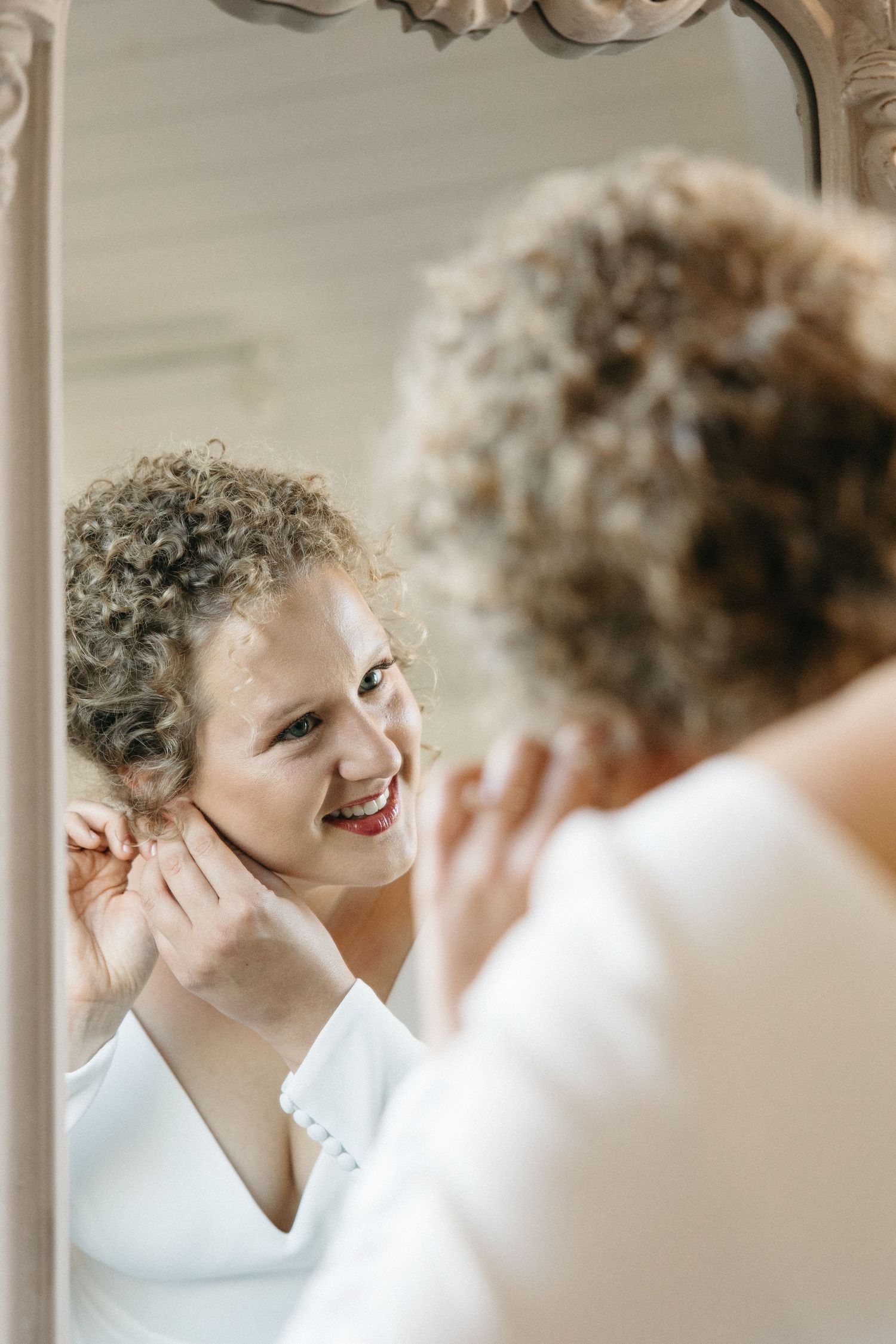 Reflection in ornate mirror shows person smiling and adjusting hair.