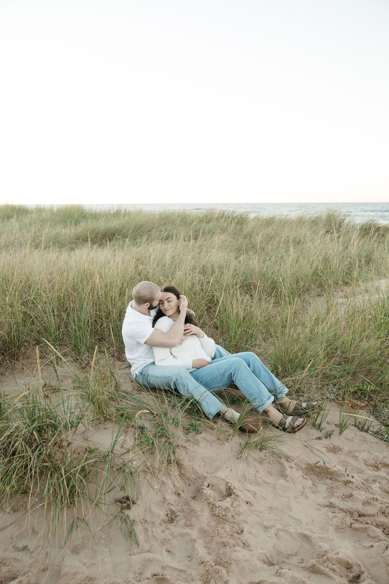 Couple cuddles on sand dunes surrounded by beach grass at dusk.