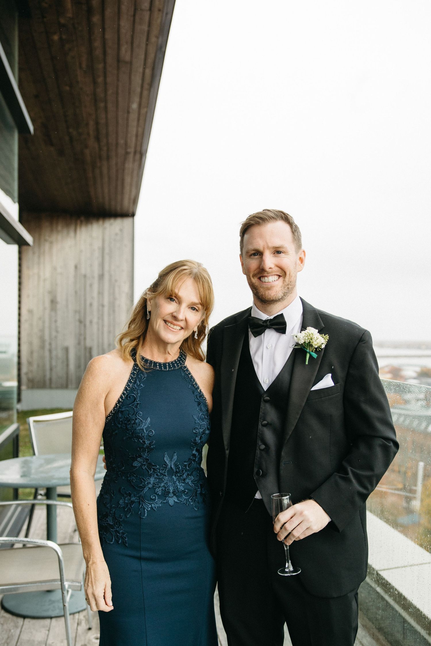 A couple in formal evening wear poses together on a modern balcony.