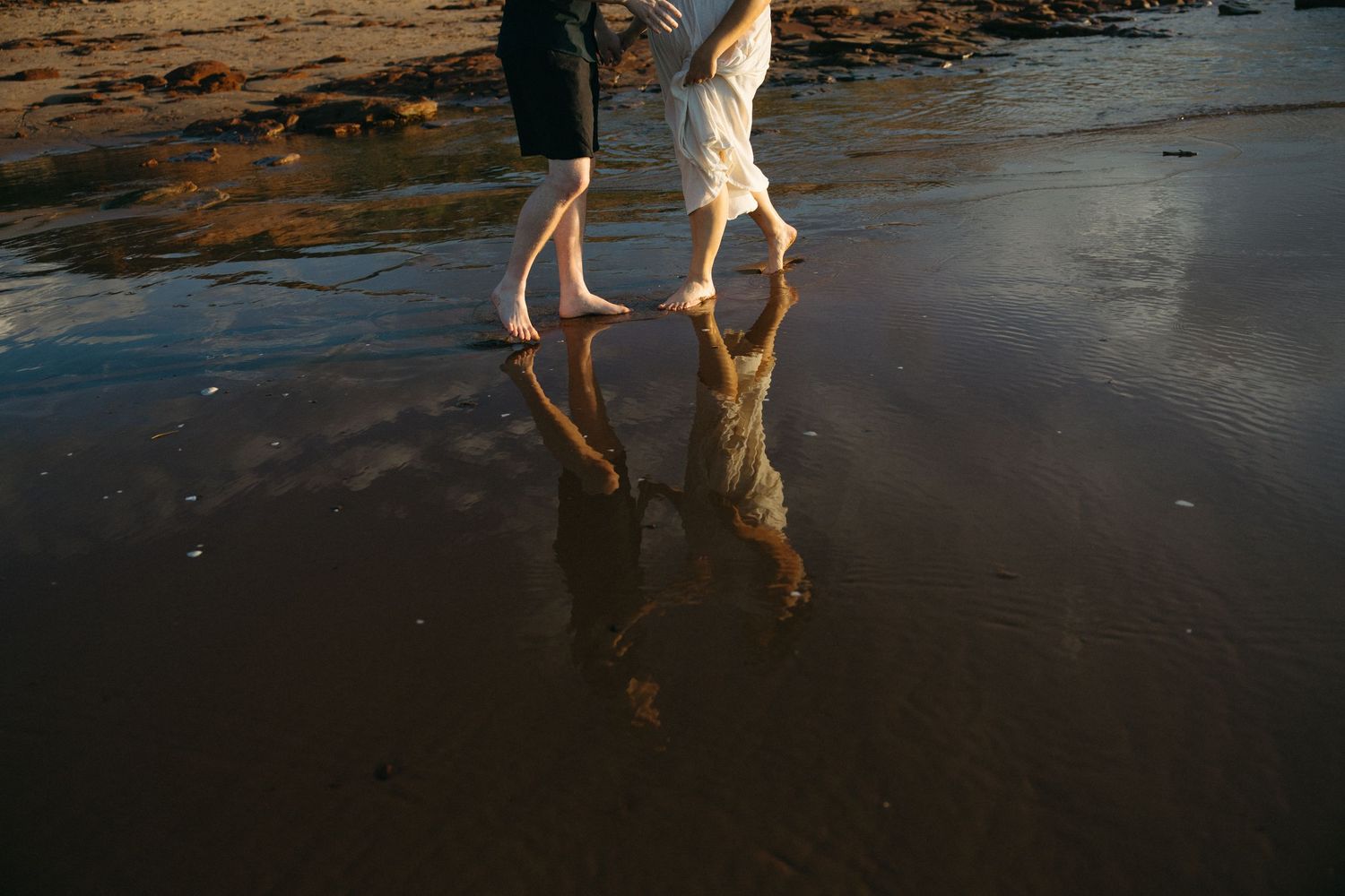 Couple walking barefoot on wet beach sand at sunset with romantic reflection in water.