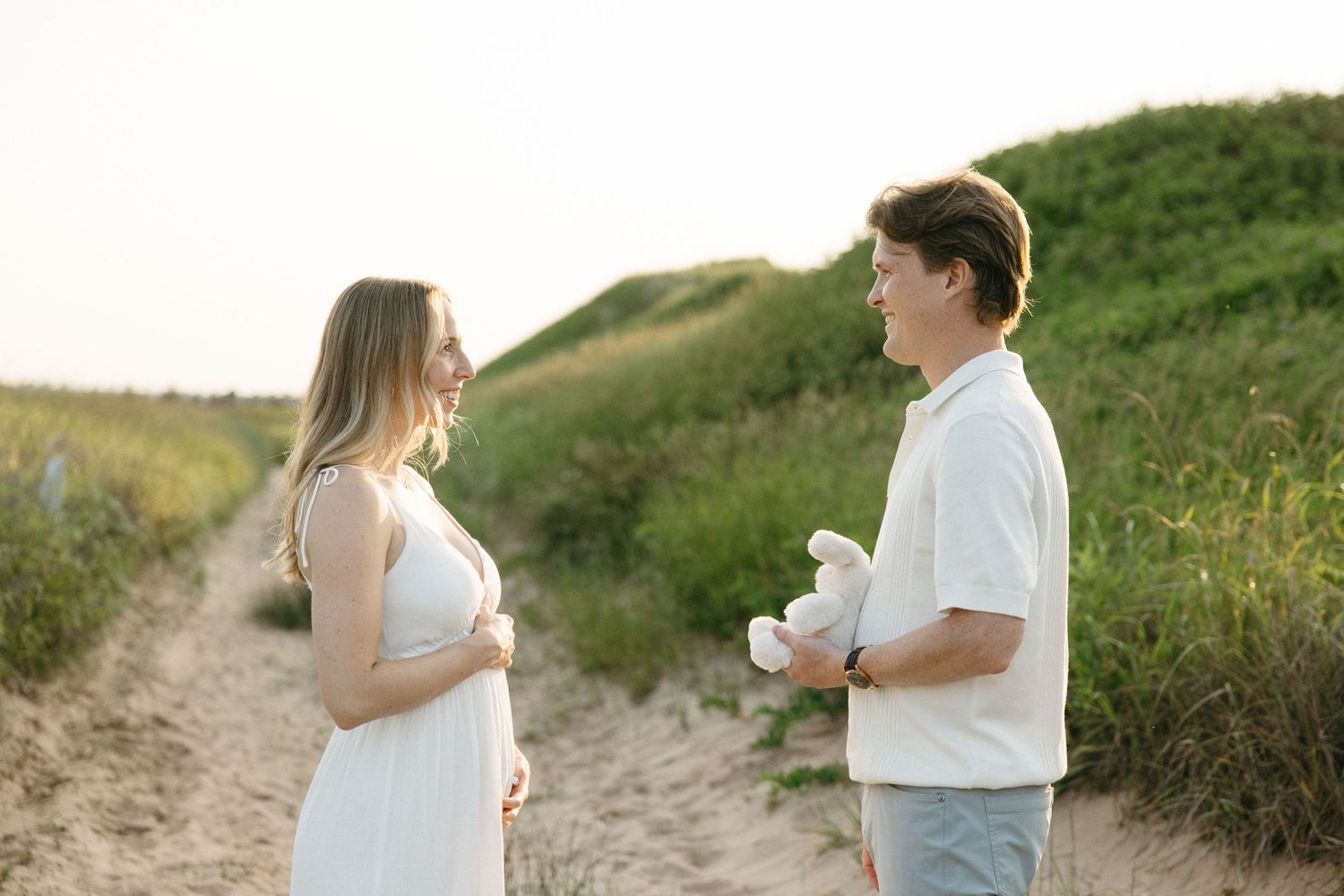 Pregnant woman in white dress and man holding stuffed animal standing on sandy path at sunset.
