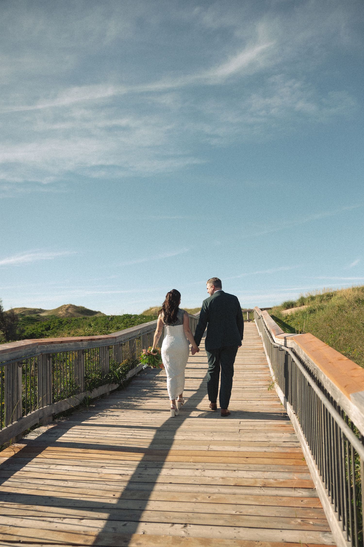 Newlyweds walk hand in hand along a wooden boardwalk toward grassy coastal dunes under a bright blue sky.