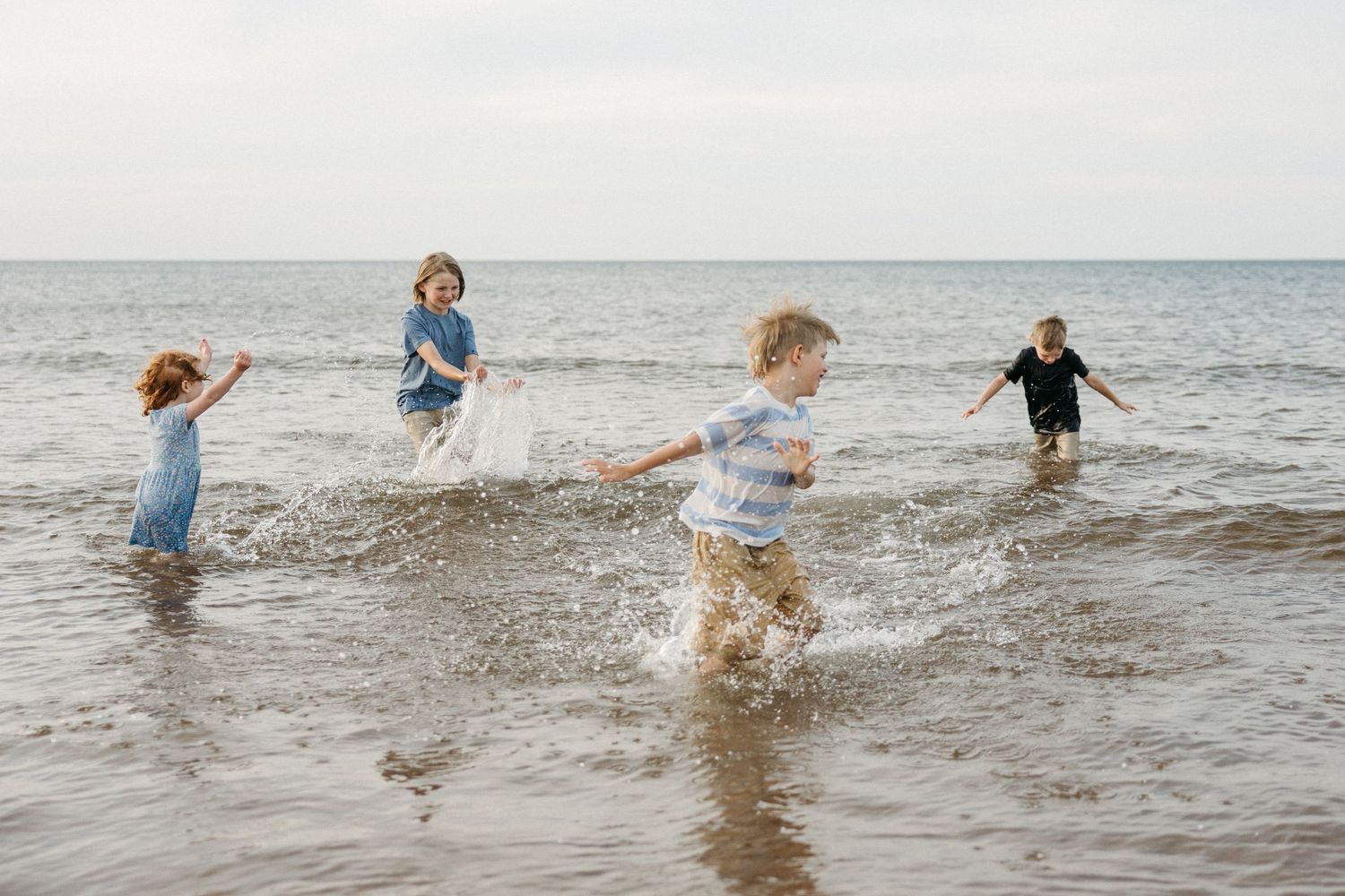 Four children splashing and playing in shallow ocean waves on a sandy beach on a cloudy day.