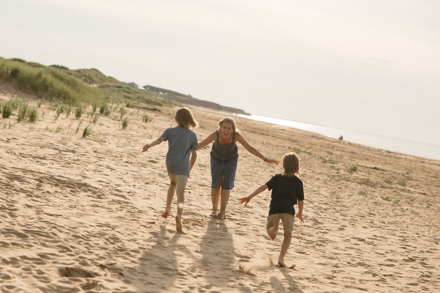 Woman holding hands with two children walking on sandy beach toward grass dunes.