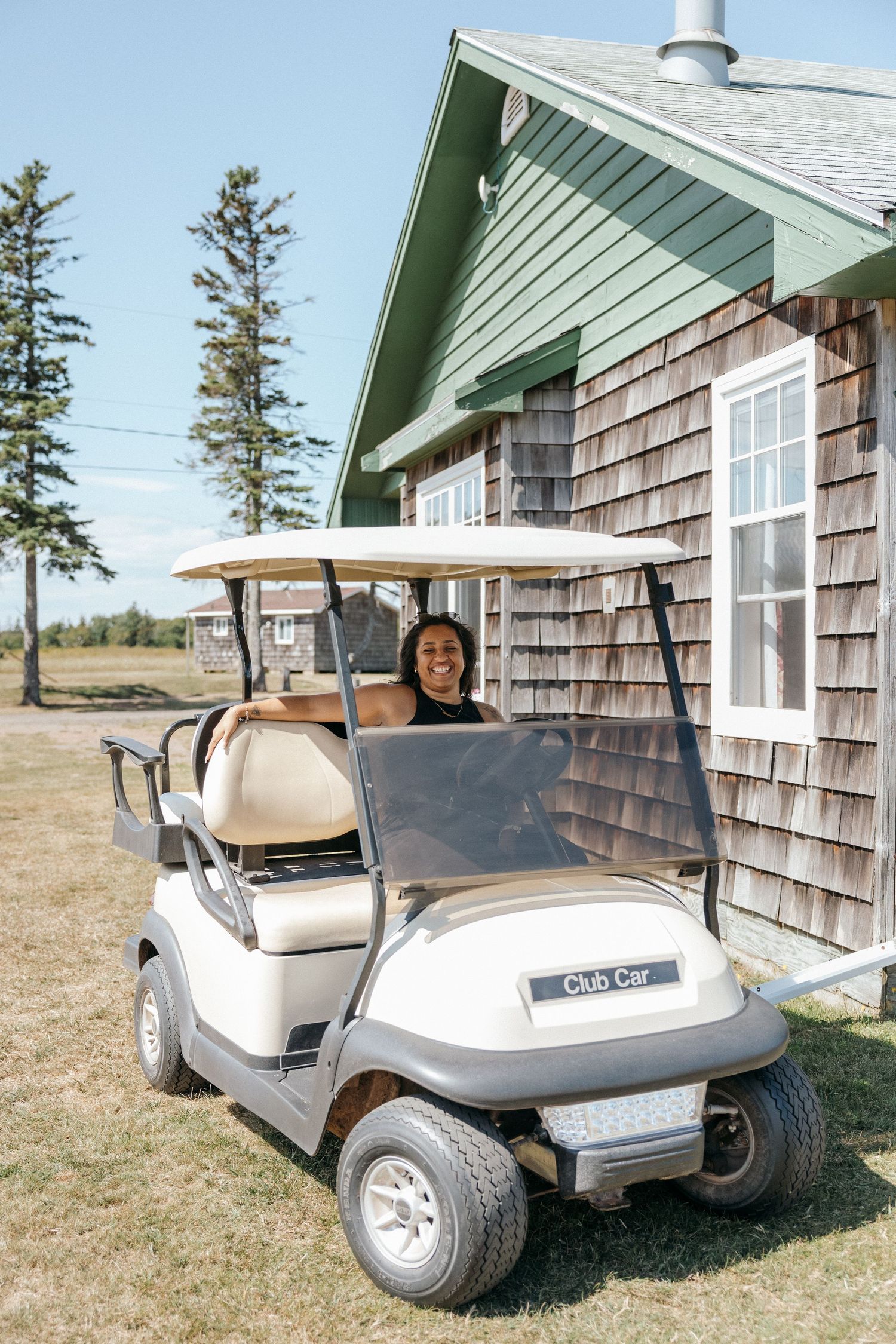 Woman smiling in a Club Car golf cart beside a cedar-shingled building on a sunny day.