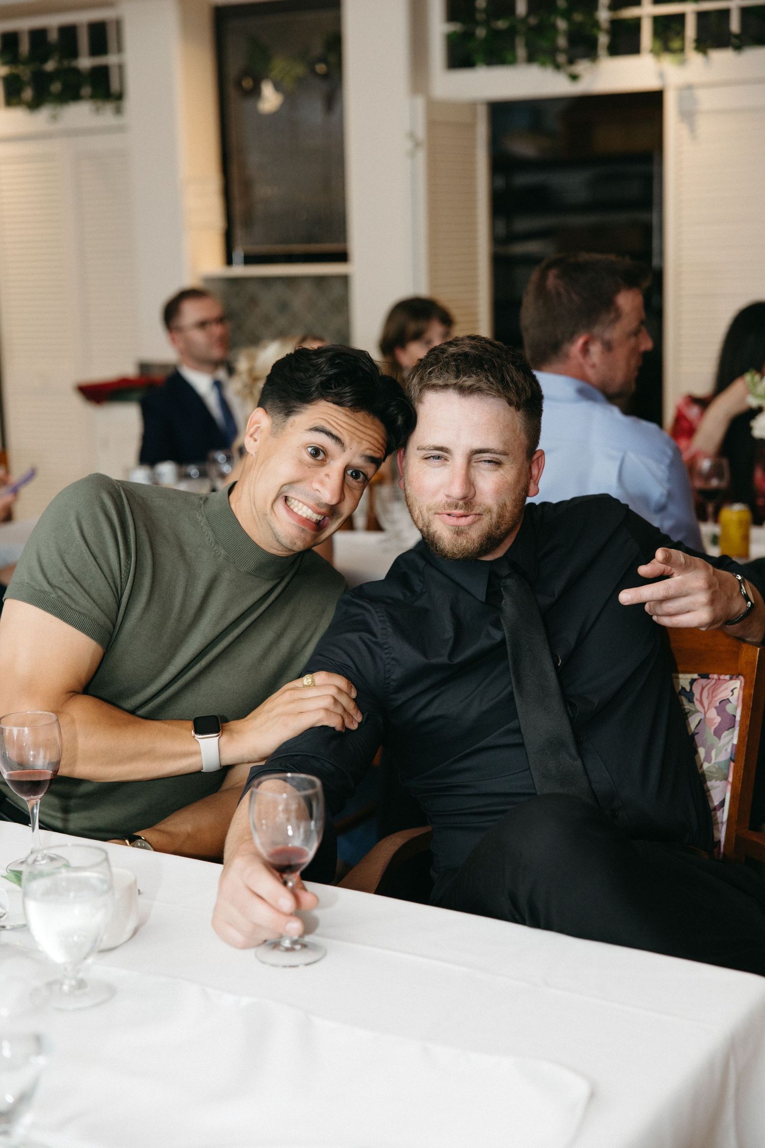 Two men smile and pose together at a wedding reception dinner table, one in olive green shirt, one in black shirt.