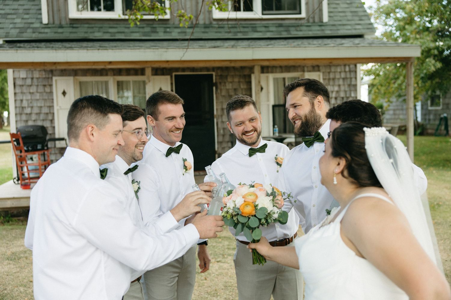 Bride in white gown and groom with groomsmen in bow ties laugh together outside rustic venue.