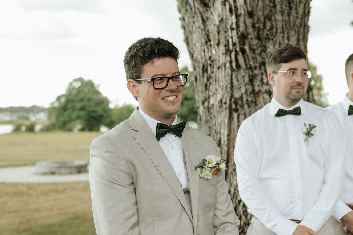 Smiling groom in gray suit and black bow tie with boutonniere stands by large tree at outdoor wedding.