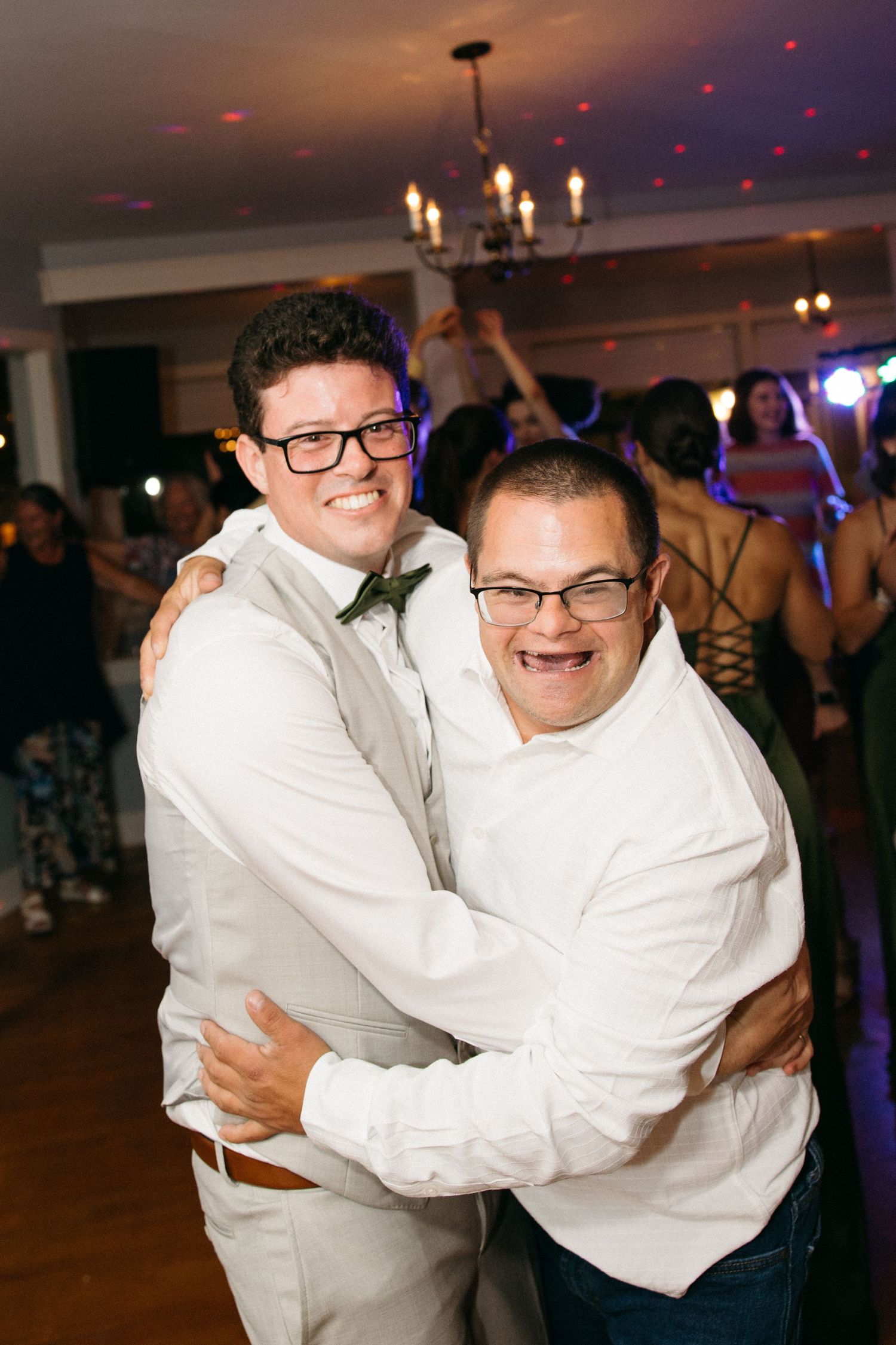 Two men in white shirts hugging and smiling on a wedding reception dance floor.