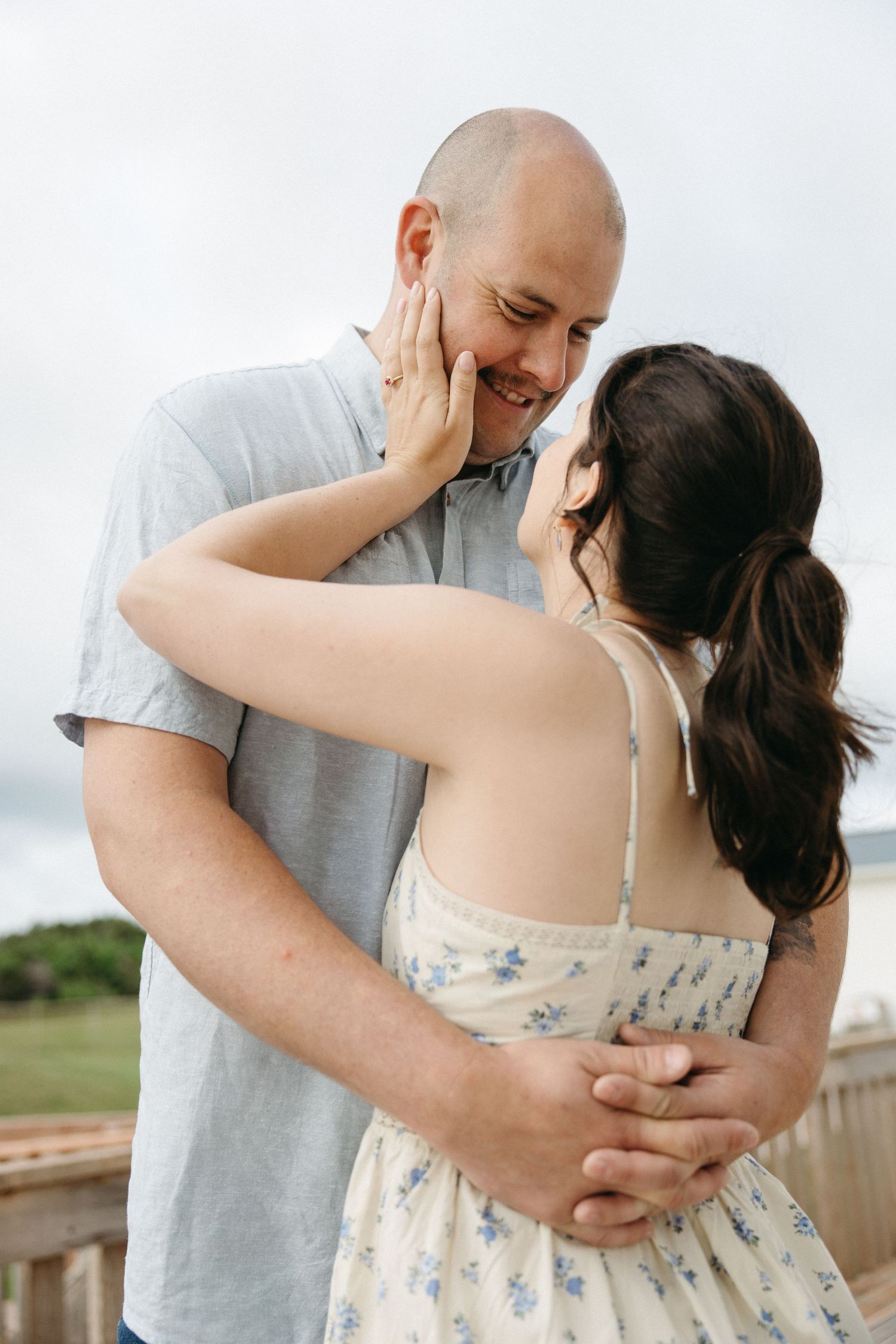Smiling couple embracing outdoors, woman in floral dress touching mans face on a cloudy day.