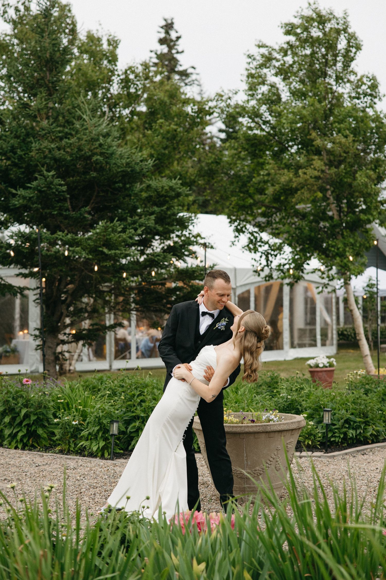 Bride and groom share a romantic dip kiss in a lush garden, with a white tent reception venue in the background.