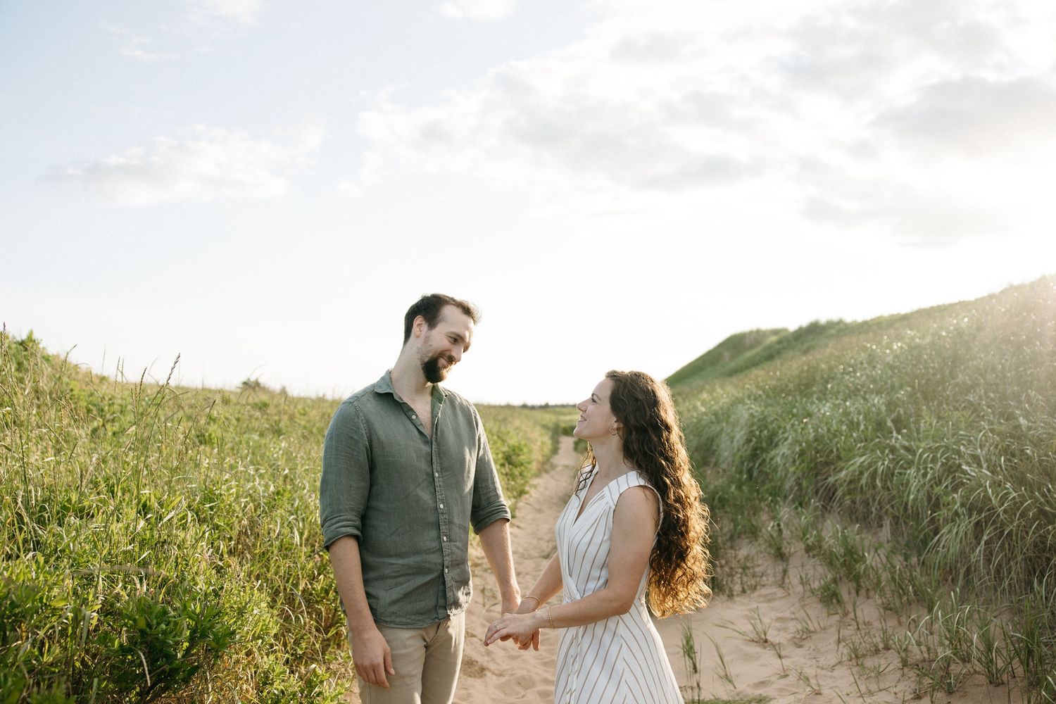 Couple holding hands on a sandy beach path surrounded by green dunes under a bright sky.