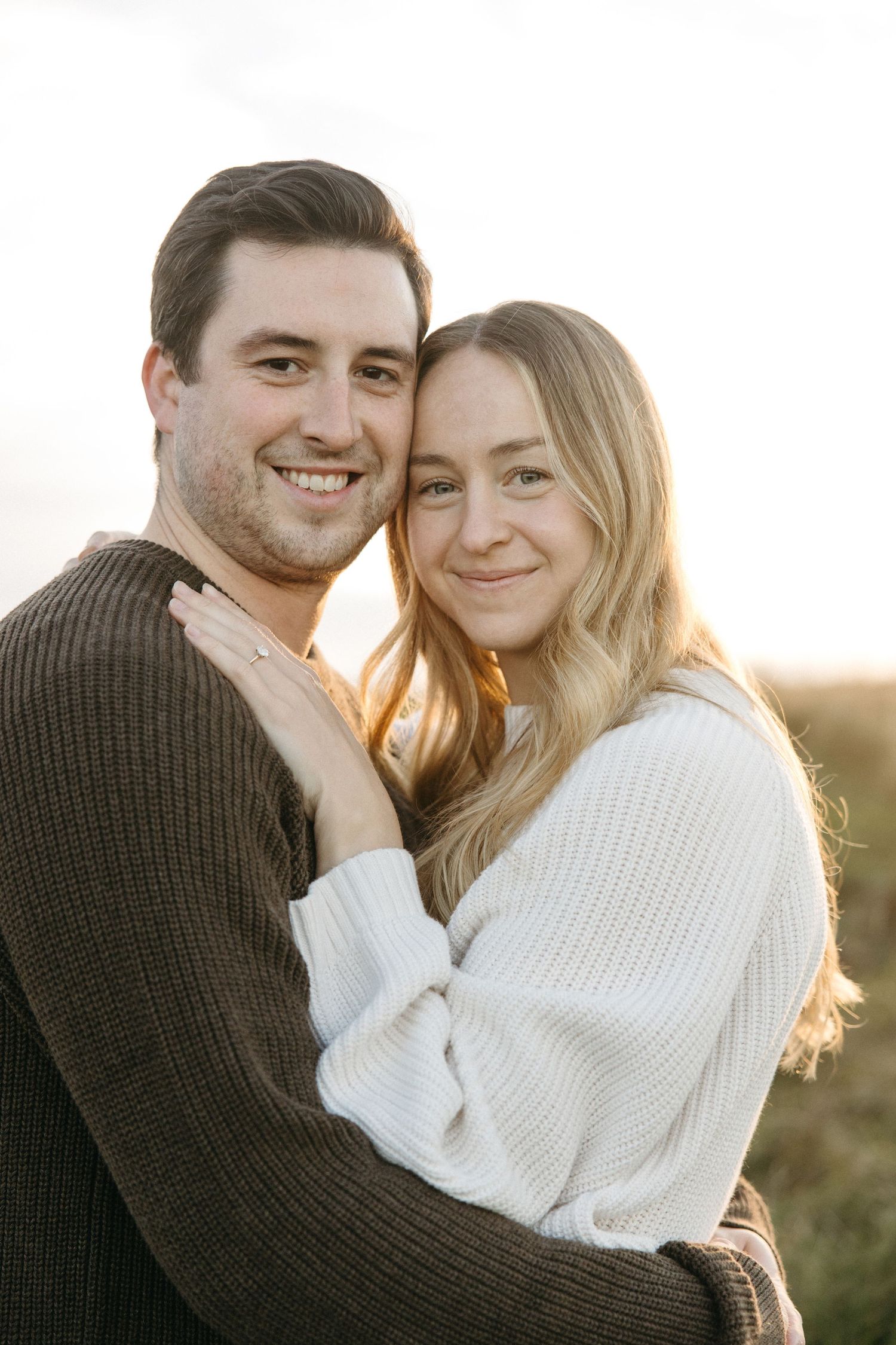 Smiling couple embrace in a grassy field at golden hour, the woman in a cream sweater showing her engagement ring beside a man in brown.