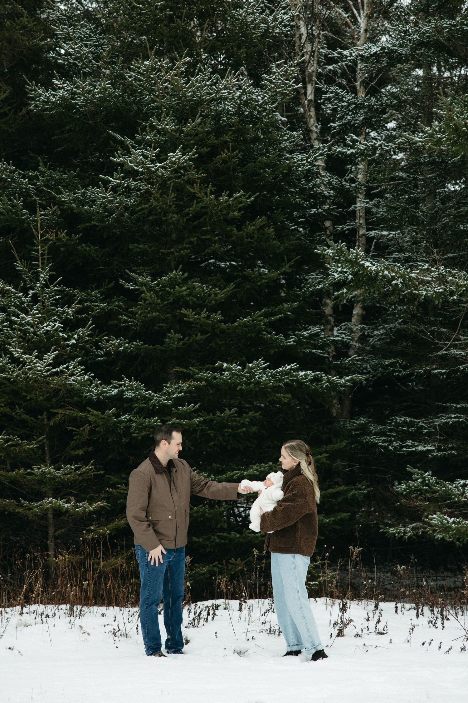 A couple shares a moment with their infant in a snow-covered forest setting.