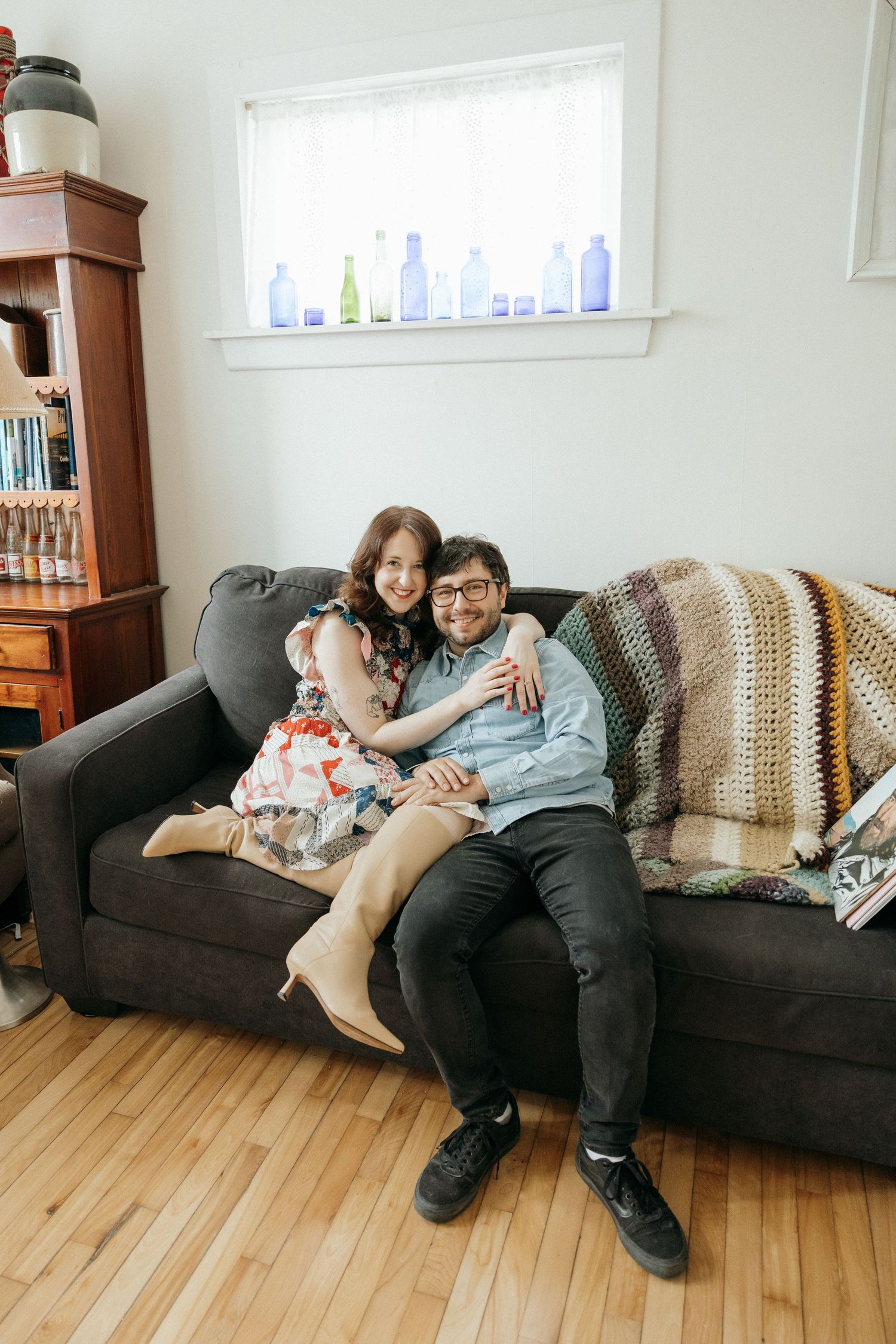 A couple lounges on a dark couch with their pets in a cozy living room setting.