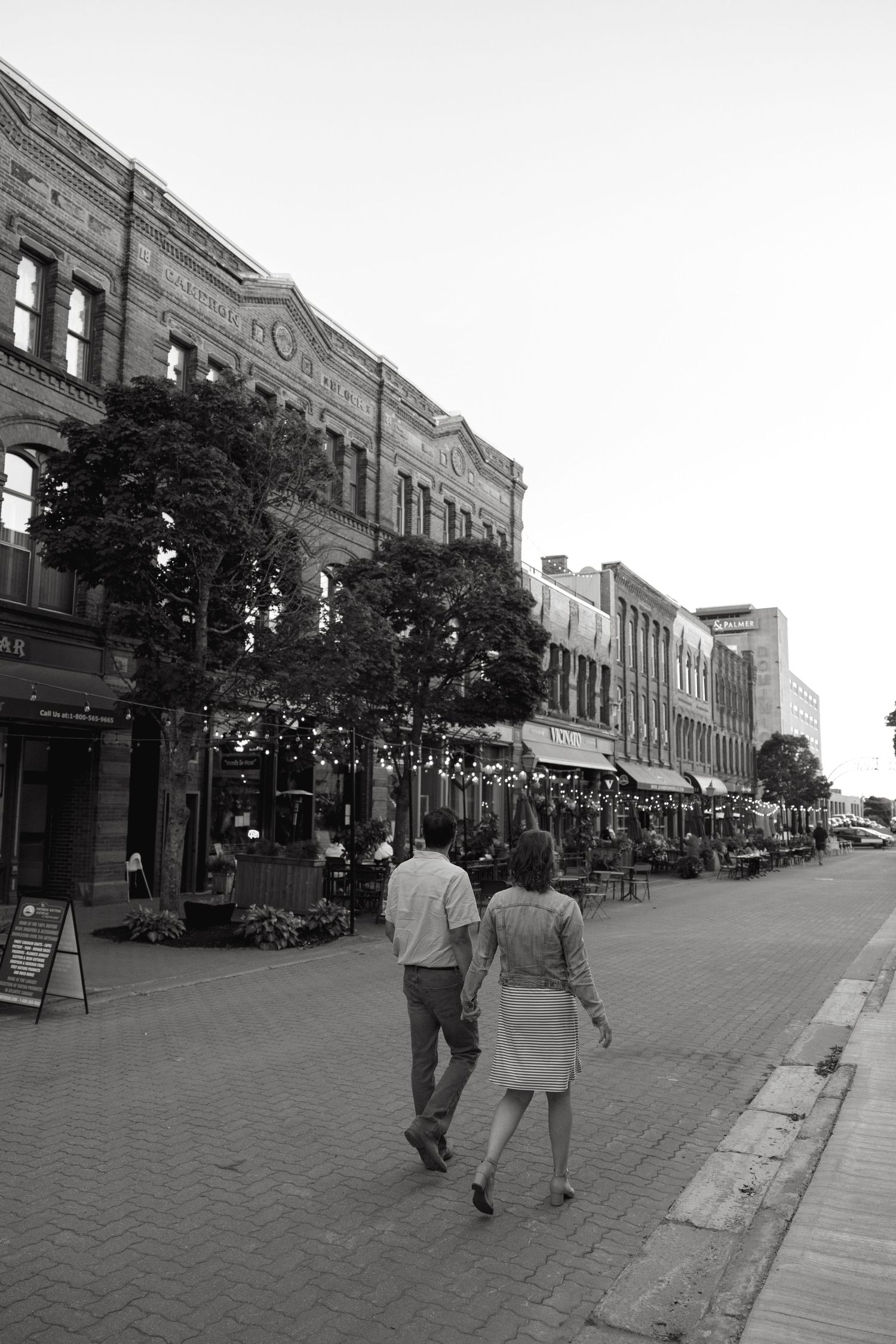 A couple strolls down a historic brick-lined street with vintage buildings and string lights.