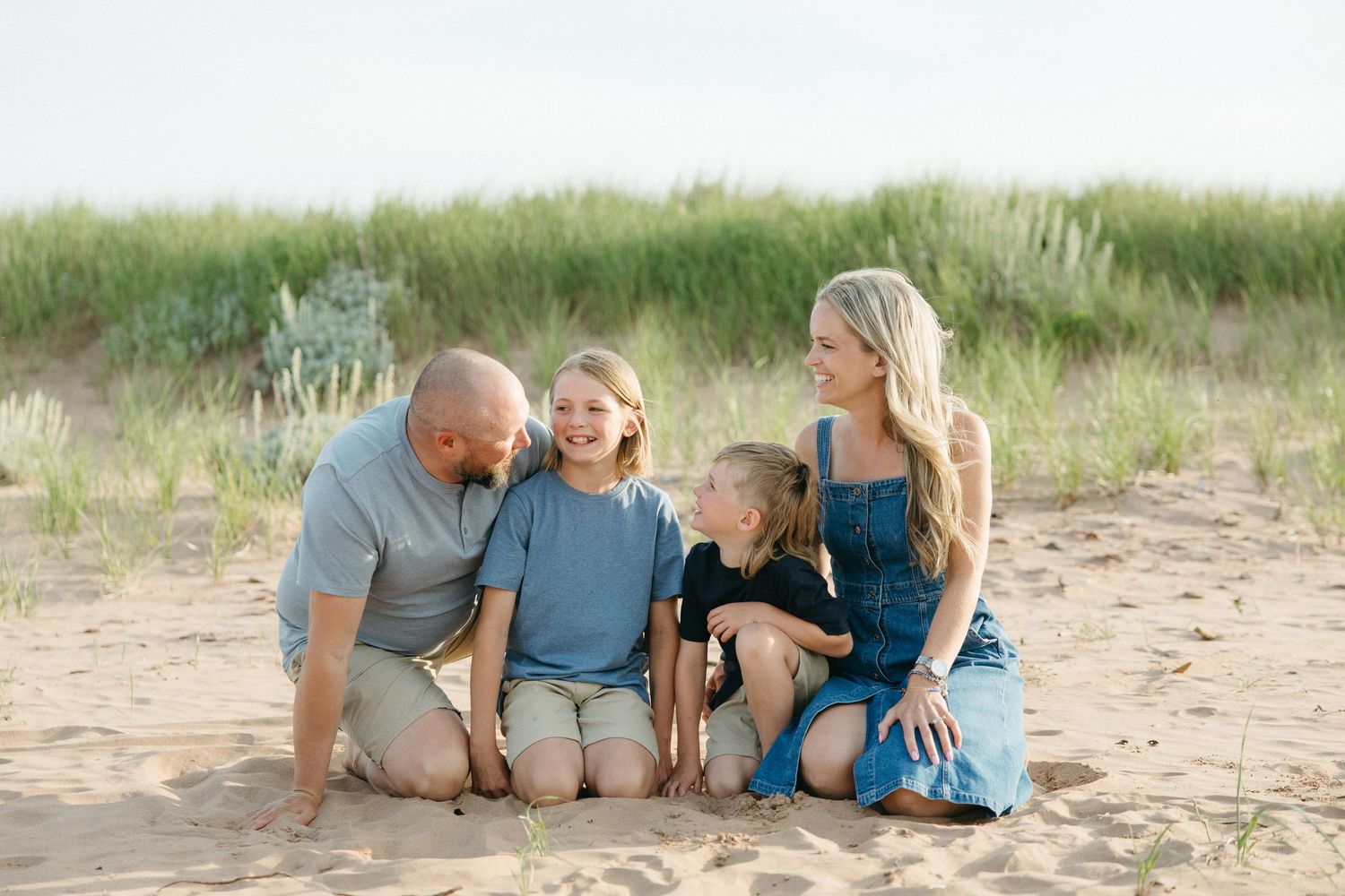 Family of four sitting together on sandy beach, smiling and laughing with green dunes in background.
