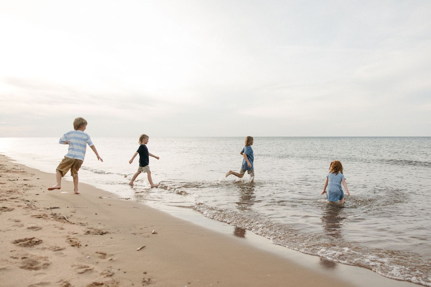 Four children running and playing along the shoreline of a sandy beach with gentle ocean waves.