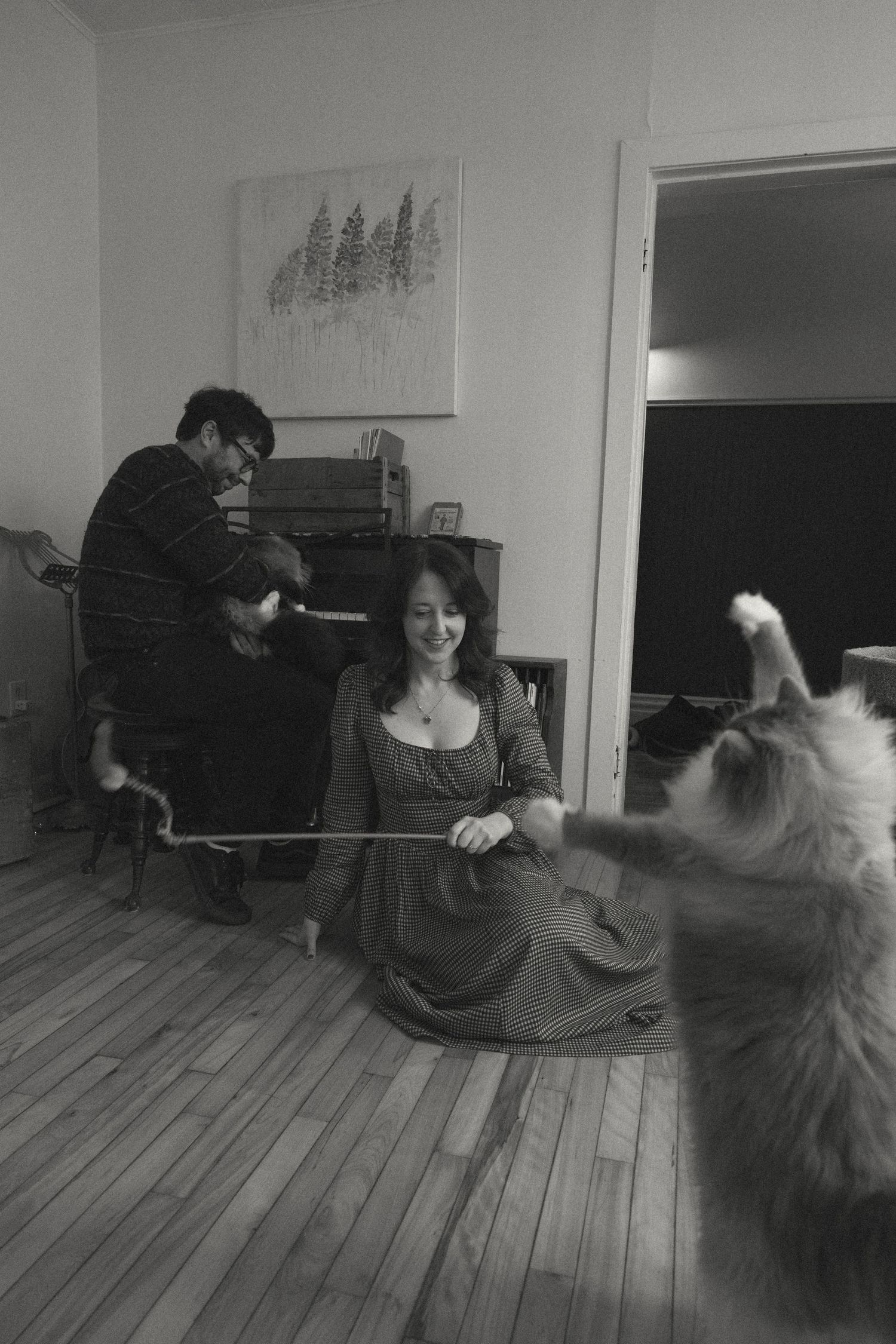 Black and white photo of a woman playing with a fluffy cat using a bow while a man and child watch near a piano.
