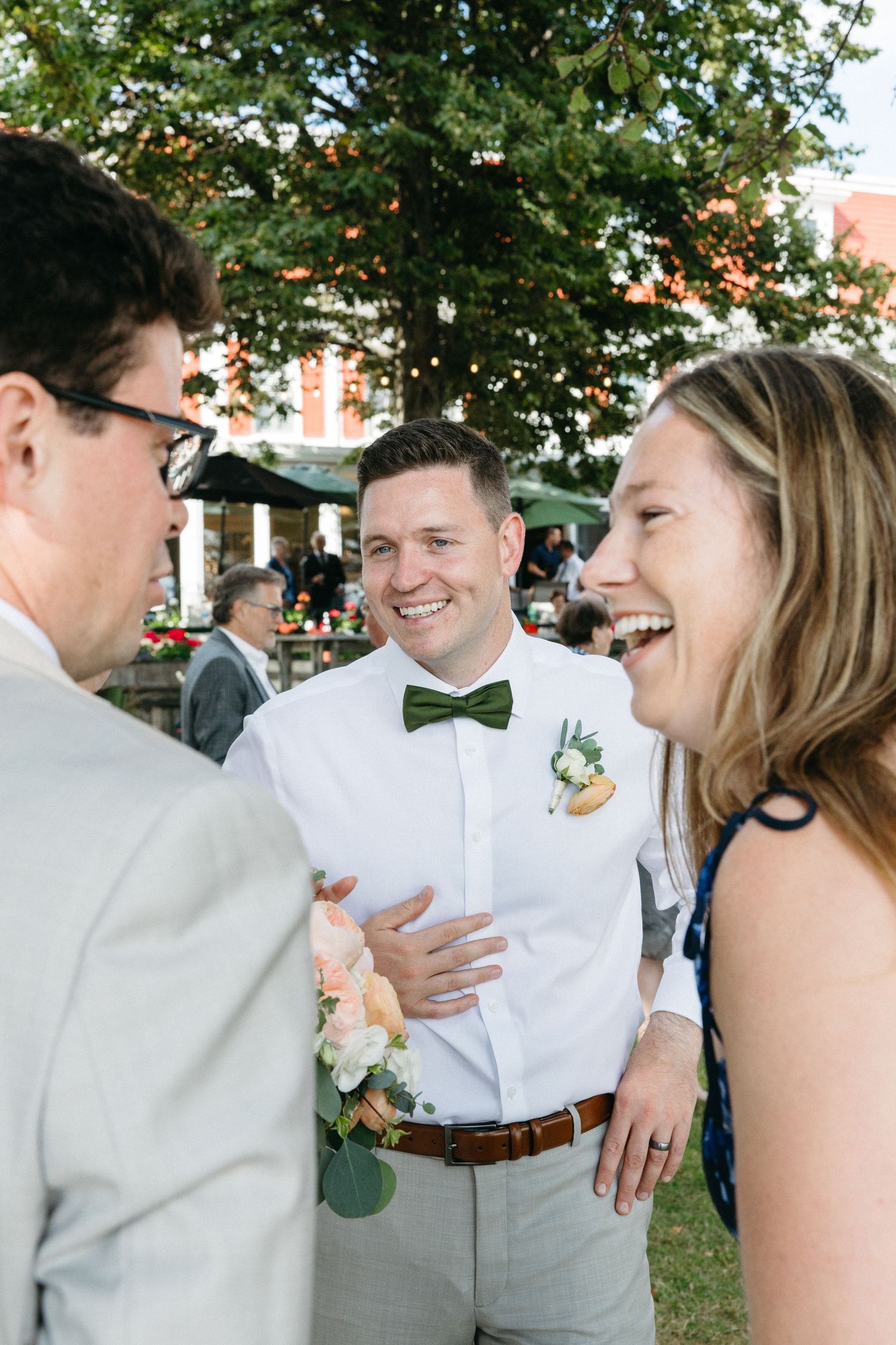 Groom in green bow tie laughing with wedding guests outdoors at summer reception.