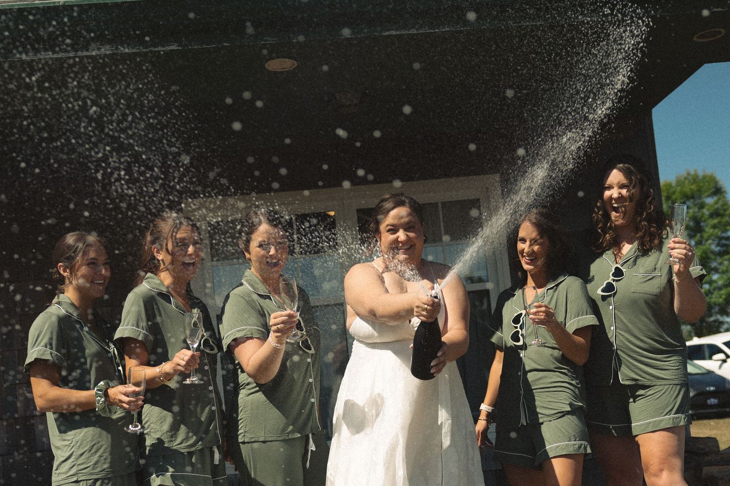 A bride in white dress pops champagne with bridesmaids in olive green dresses outdoors.