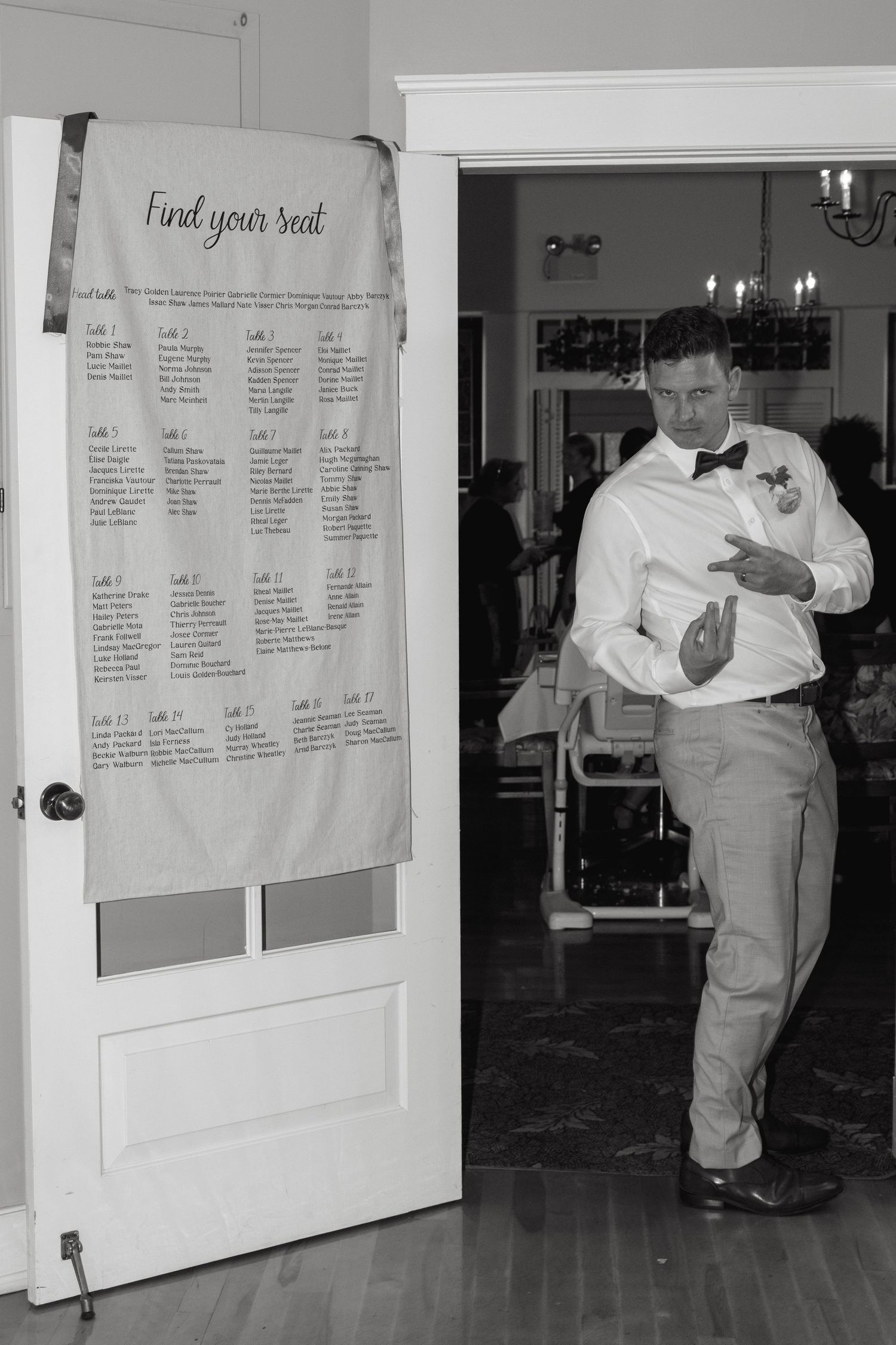 Groom in white shirt and bow tie enters reception hall beside a wedding seating chart on a door.