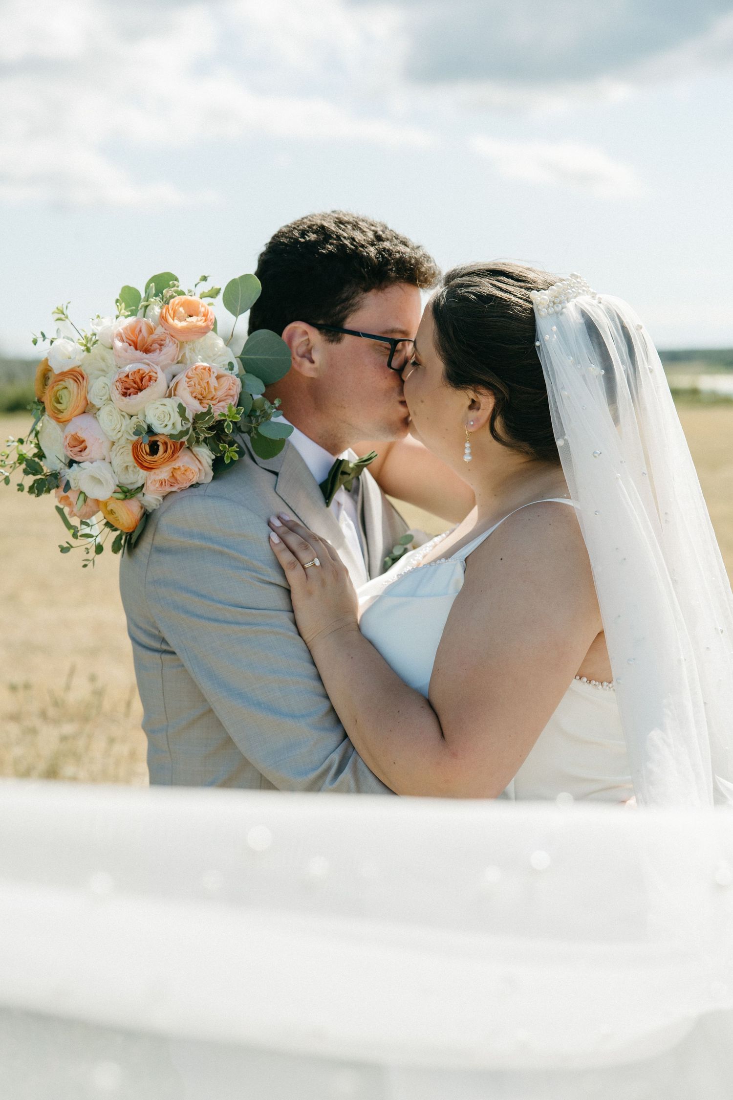 Bride and groom sharing a kiss outdoors holding a peach and white floral bouquet on their wedding day.