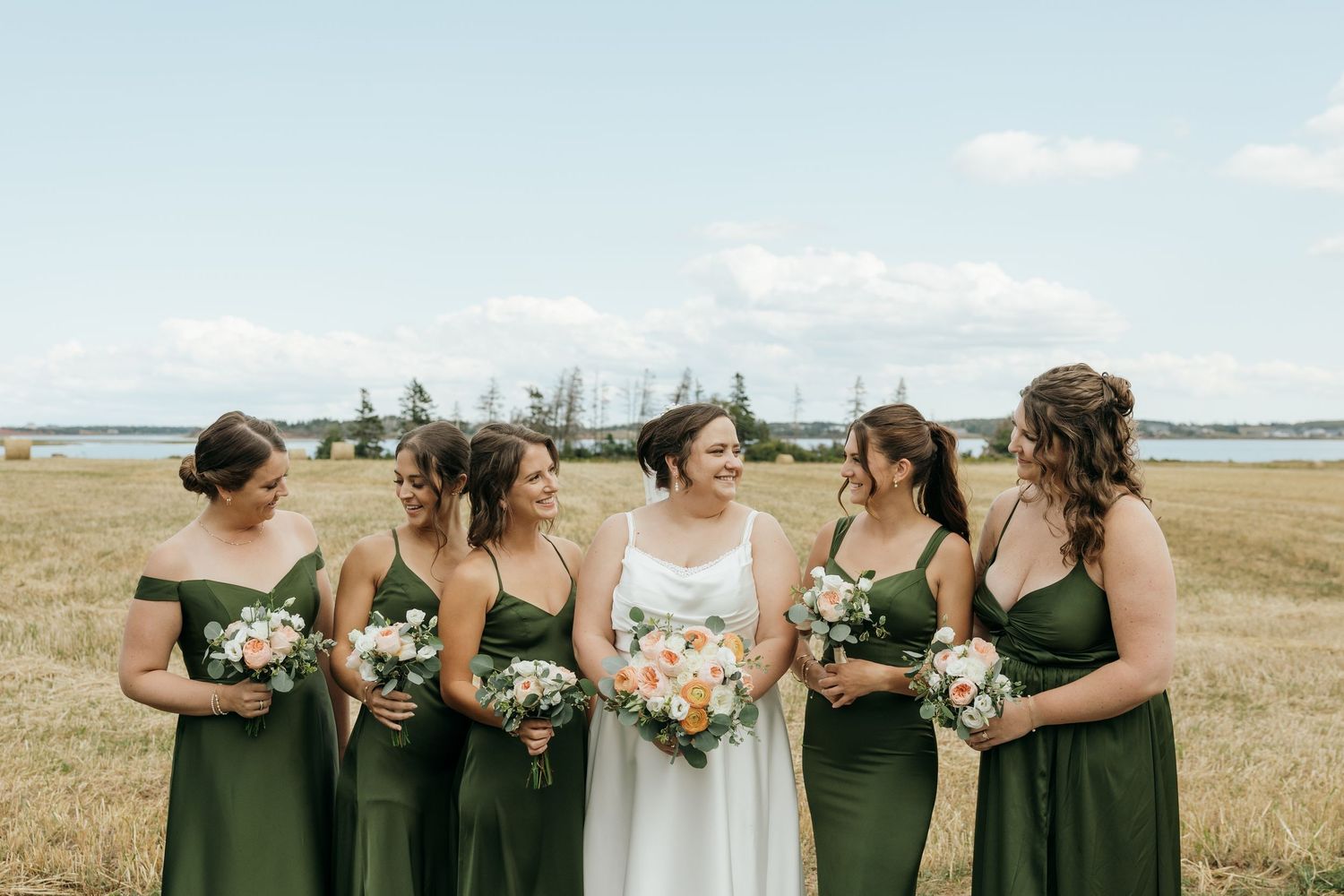 Bride in white gown with peach bouquet laughing with five bridesmaids in olive green dresses in a golden field.