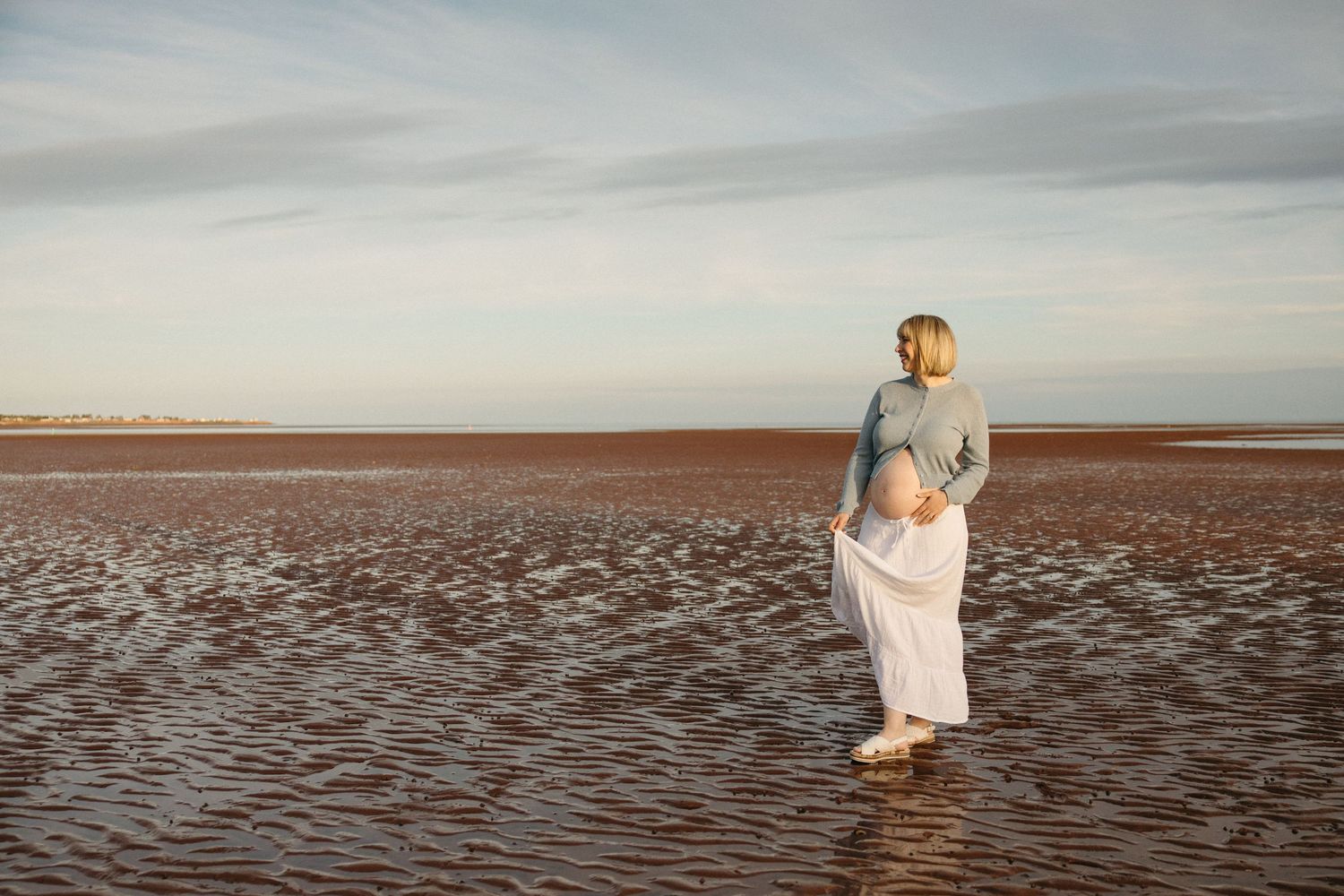 Pregnant woman standing barefoot on a wet sandy beach, wearing a white skirt and gray top, gazing into the distance.