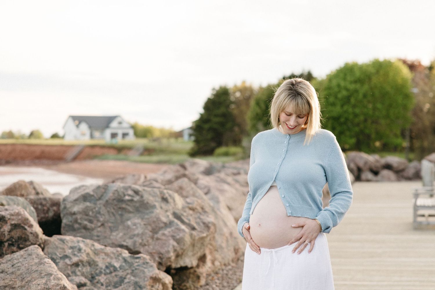 Pregnant woman in blue sweater and white skirt cradling her baby bump outdoors near rocky shoreline.